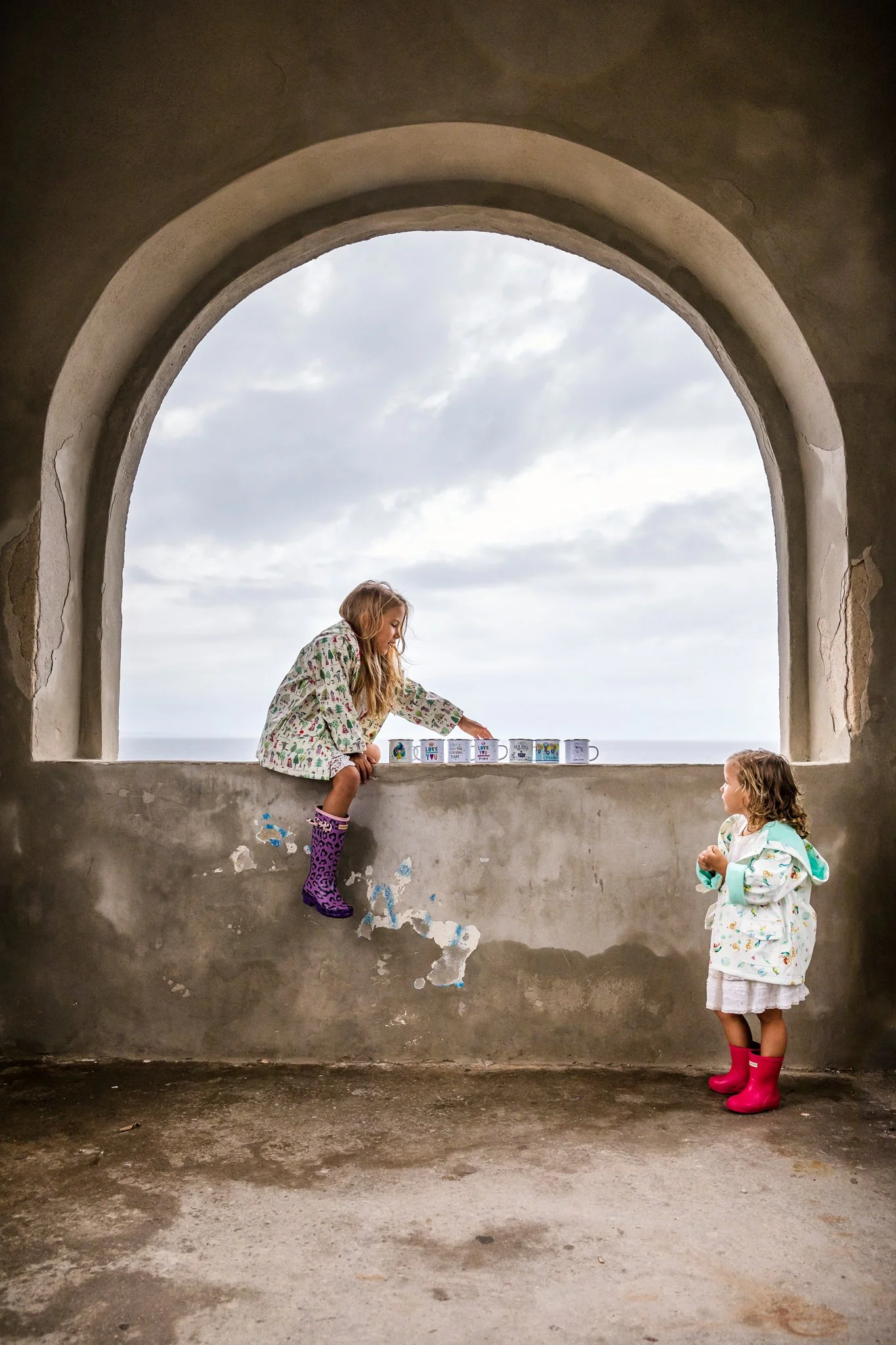 Two young girls with curly blonde hair are playing in front of a large arched window. One girl is standing on the window ledge, reaching out to the other girl, who is standing on the ground. The girl on the ledge is wearing a colorful dress and purpl