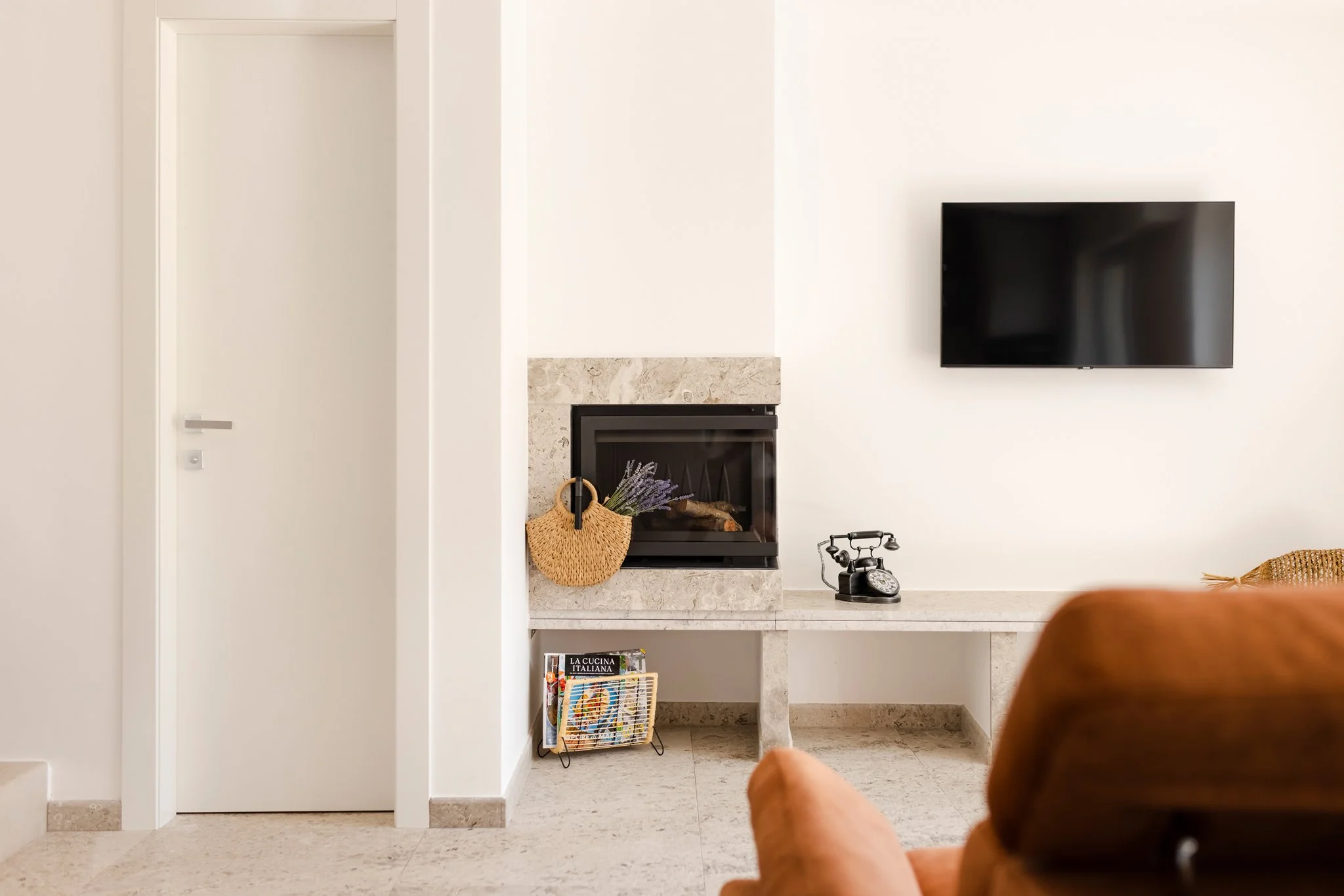 A living room corner featuring a fireplace with a beige stone surround, a black flat-screen TV mounted on a white wall, a woven basket with lavender flowers hanging on the side of the fireplace, a vintage black rotary phone on a marble ledge, a magaz