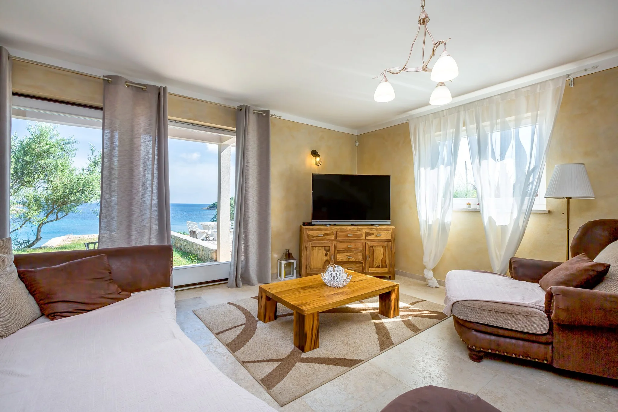 A living room with large windows showing an ocean view, beige walls, a wooden coffee table, a flat-screen TV on a wooden stand, a white sofa with brown pillows, a floor lamp, and curtains letting in sunlight.