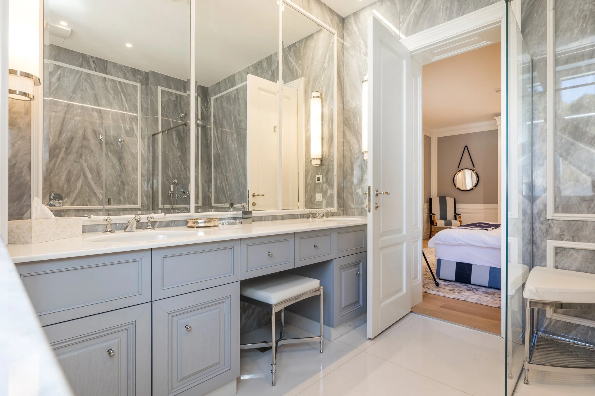 A luxurious bathroom with marble walls, a large mirror, and a white vanity with multiple drawers. There is a small stool beneath the vanity. The reflection shows part of a doorway leading to a bedroom with a bed and a mirror hanging on the wall. The 