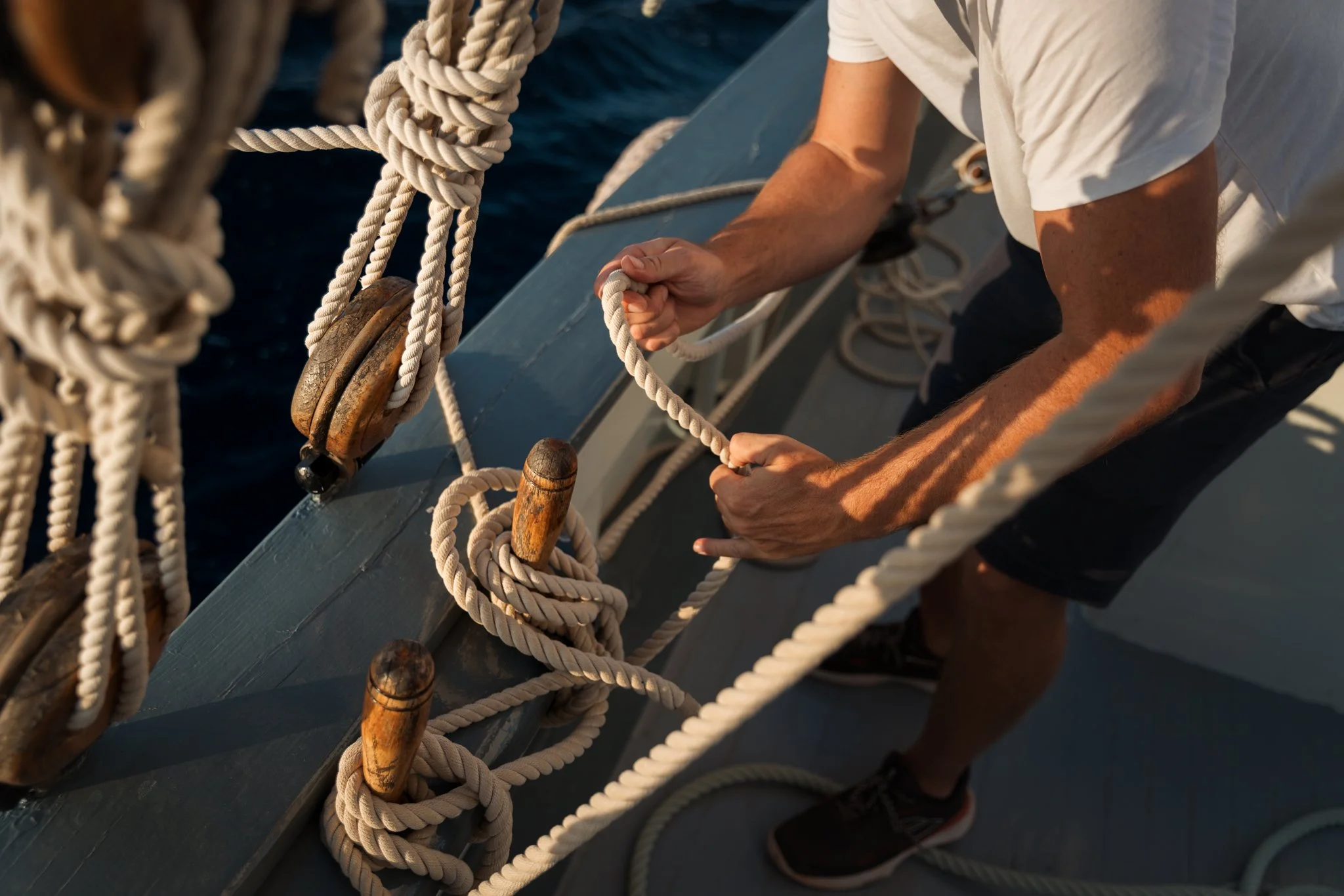 Close-up of a person's hands pulling on a thick white rope tied around wooden pulleys on a boat deck. The person is wearing a white shirt and black shorts. The background shows the dark blue water.