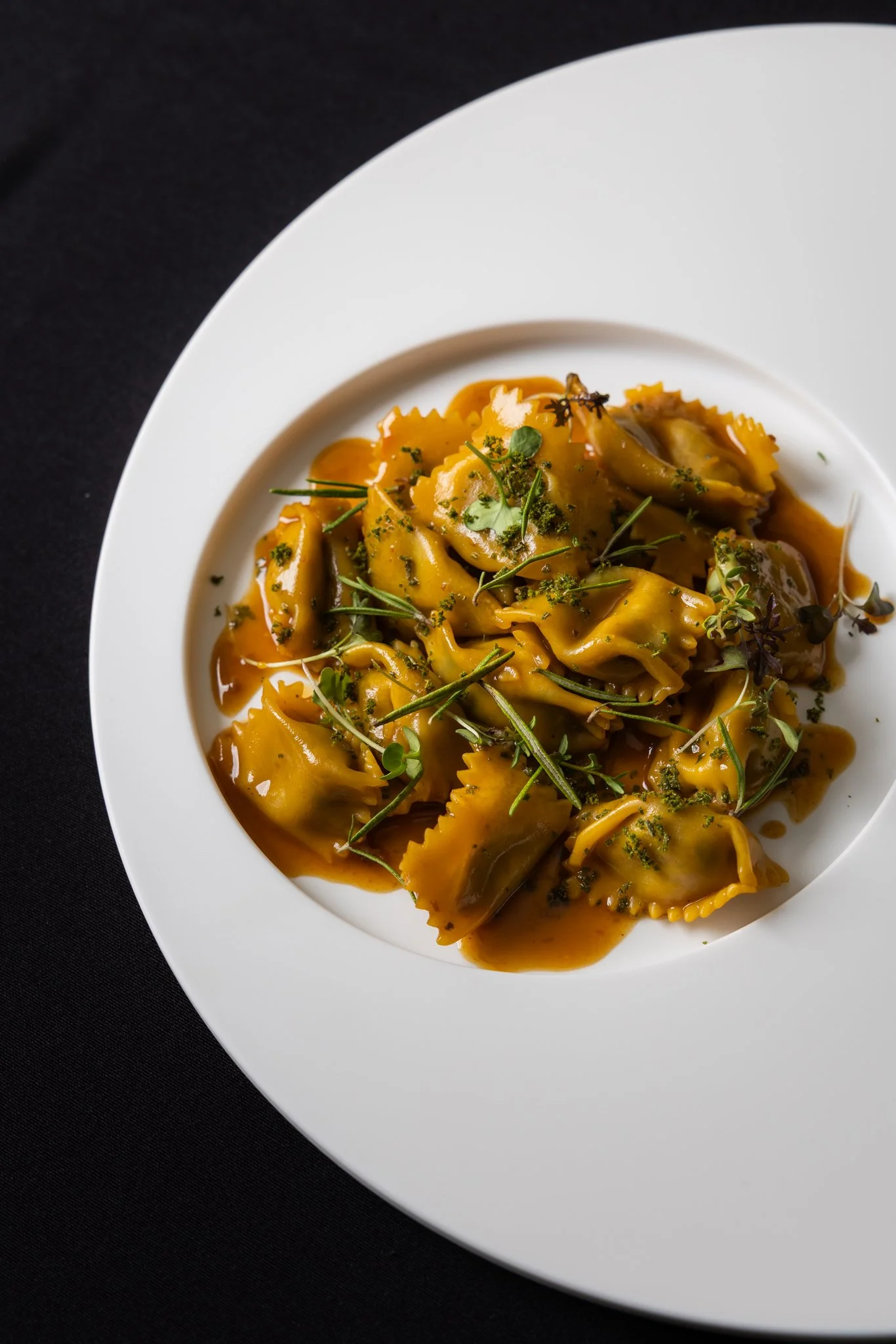 Close-up image of a white plate of ravioli pasta with brown sauce, garnished with herbs and microgreens, on a black surface.