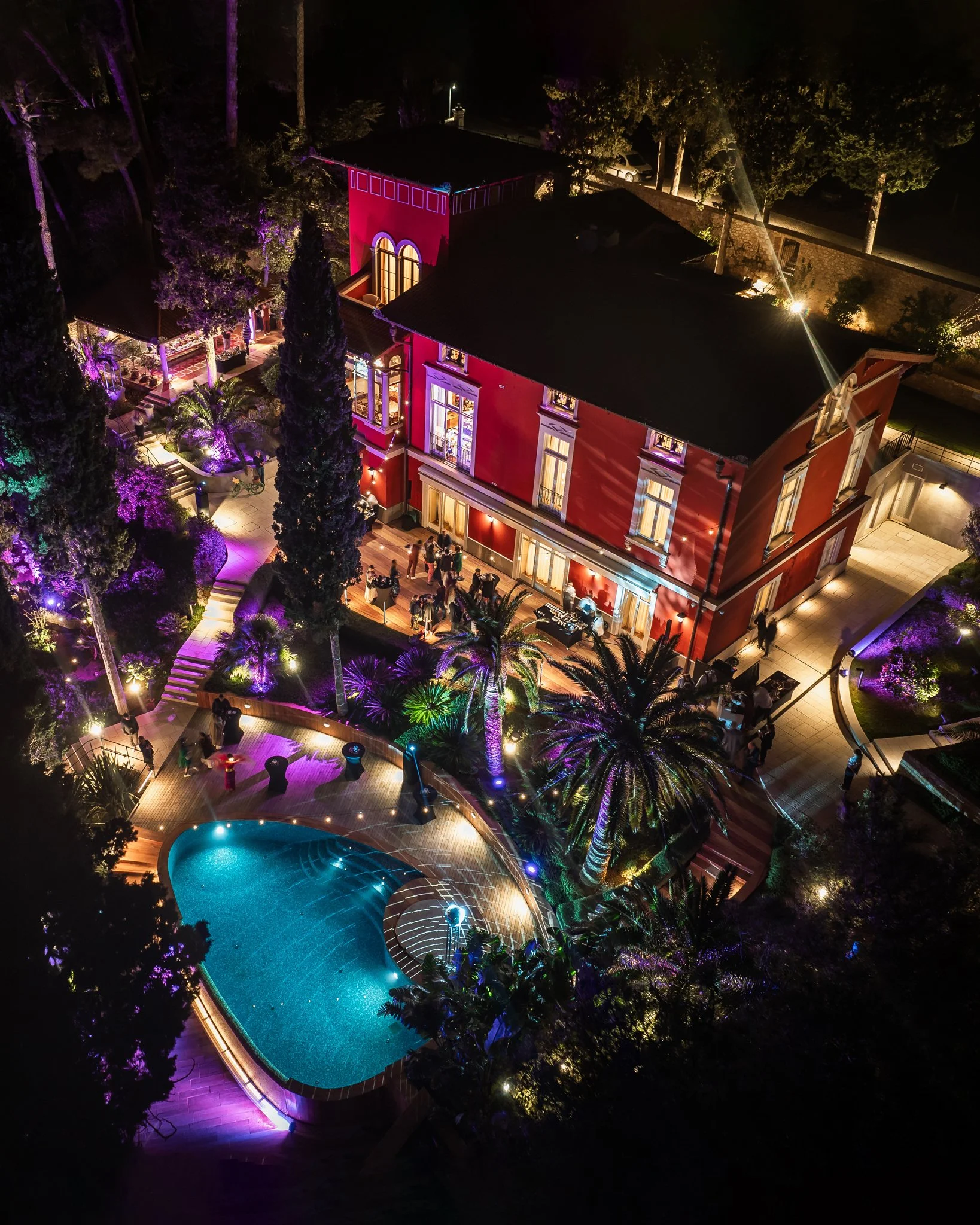 Nighttime aerial view of a brightly lit backyard with a swimming pool, surrounded by lush trees, plants, and a red house with large windows and outdoor lighting.