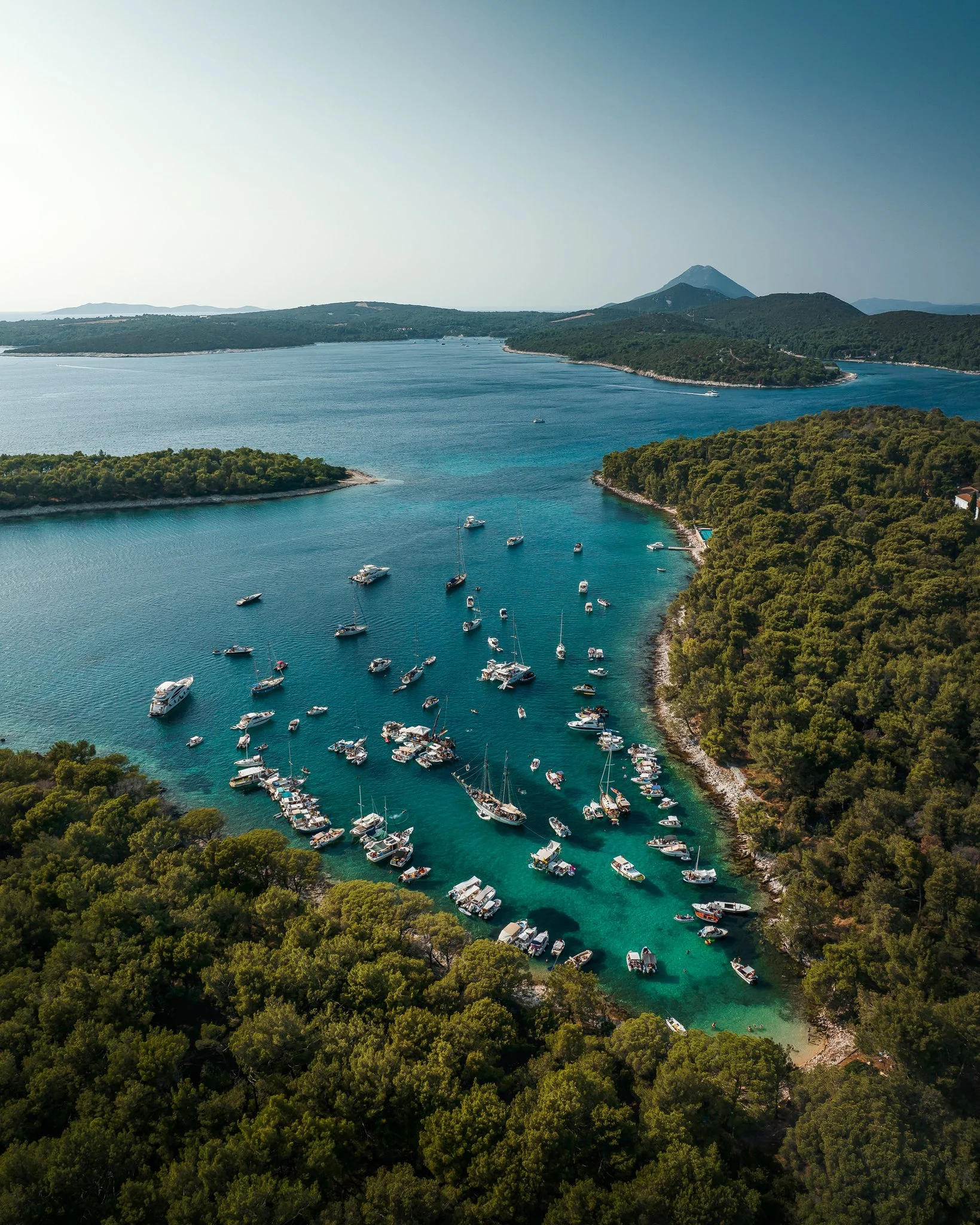 Aerial view of a harbor with numerous boats and yachts anchored in clear blue water, surrounded by green wooded islands and hills, with a mountain in the background under a clear sky.