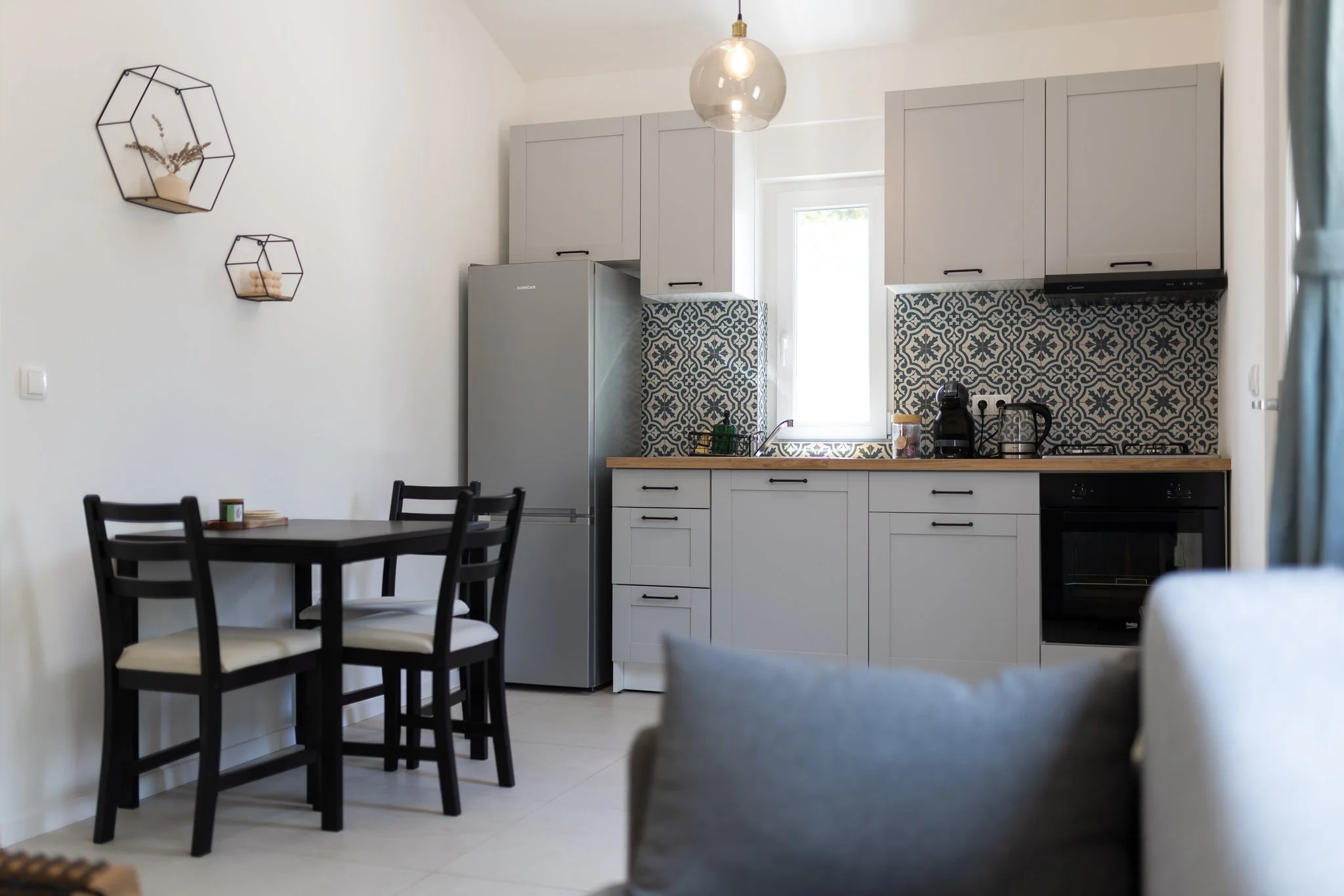 A modern kitchen with white cabinets, black hardware, a gray refrigerator, and blue patterned tile backsplash under a window. There is a wooden countertop, black coffee maker, and black stove. A black dining table with four chairs is in the foregroun
