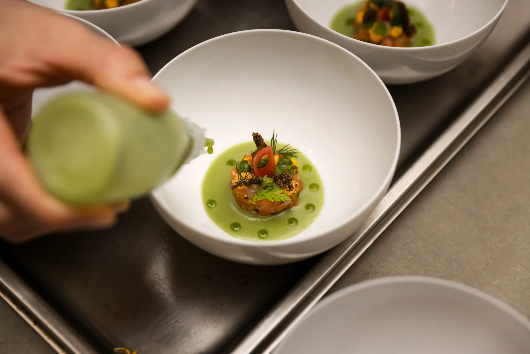 Chef drizzling green sauce over an artfully arranged appetizer in a white bowl with other similar bowls in the background.