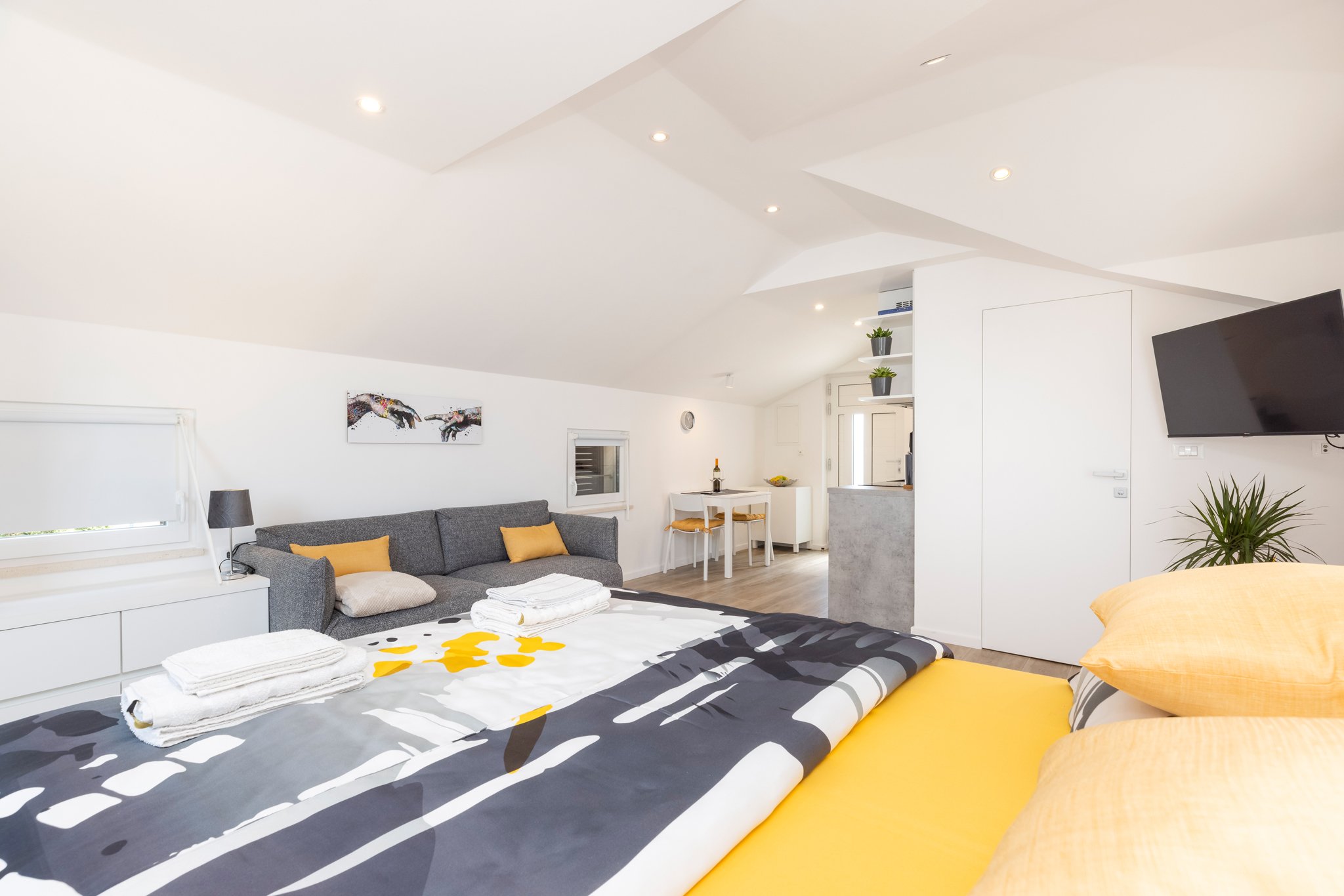 Modern, minimalistic living room with a grey sofa, yellow pillows, a black and white patterned rug with yellow accents, a bed with white linens and towels, a wall art depicting hands reaching, and a mounted flat-screen TV.