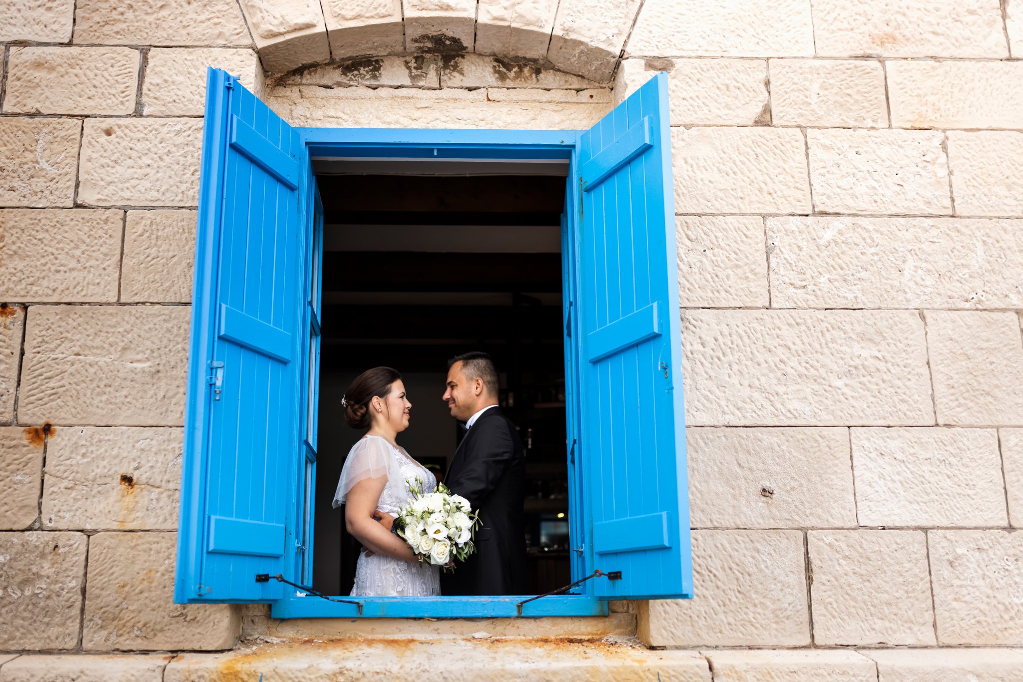 A bride and groom facing each other inside a window with bright blue shutters, set in a stone wall. The bride wears a white wedding dress and holds a bouquet of white flowers, while the groom wears a black tuxedo with a bow tie.