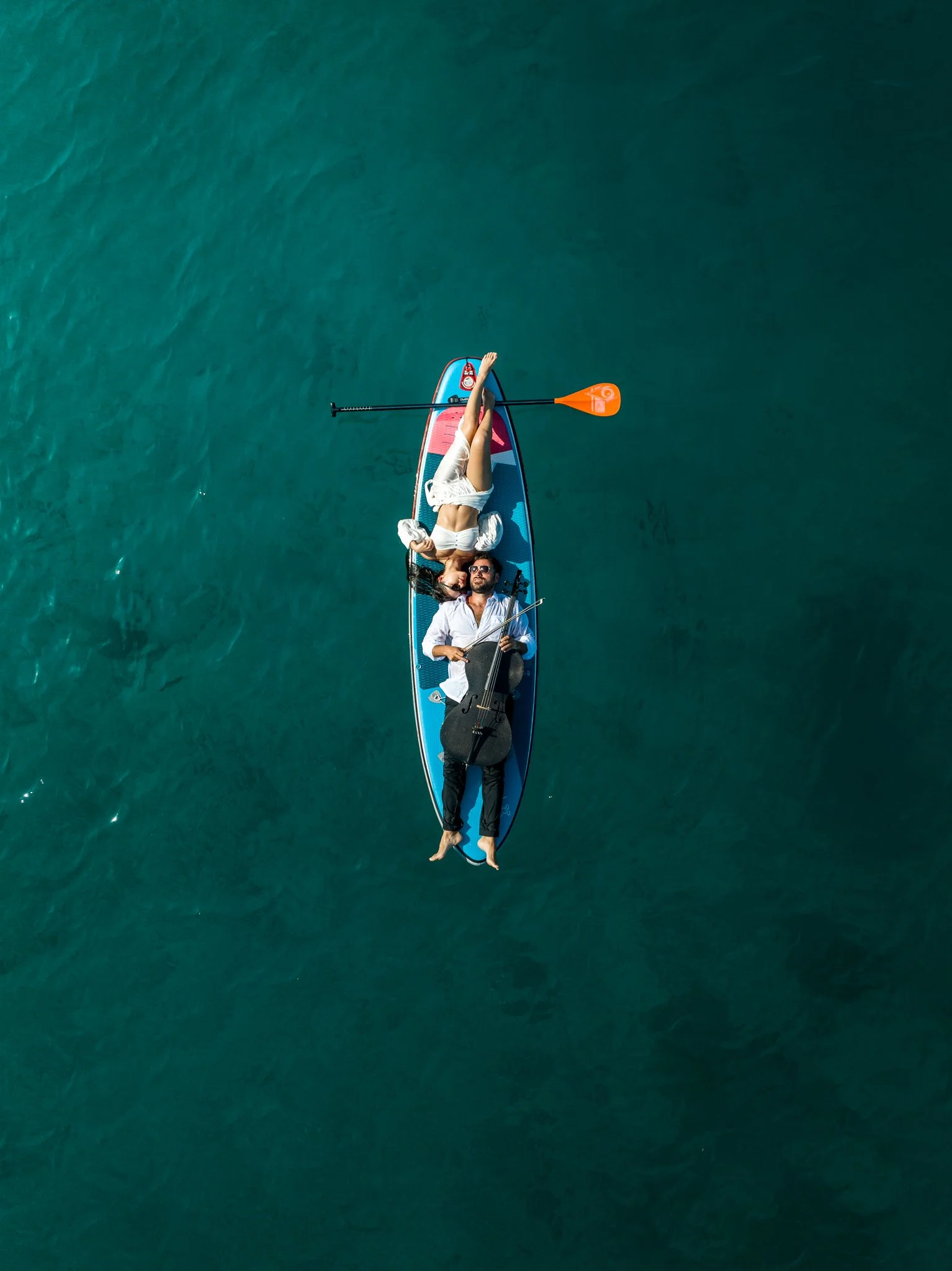 A couple lying on a stand-up paddleboard in the water, with the woman resting her head on the man's chest and the man holding a cello.