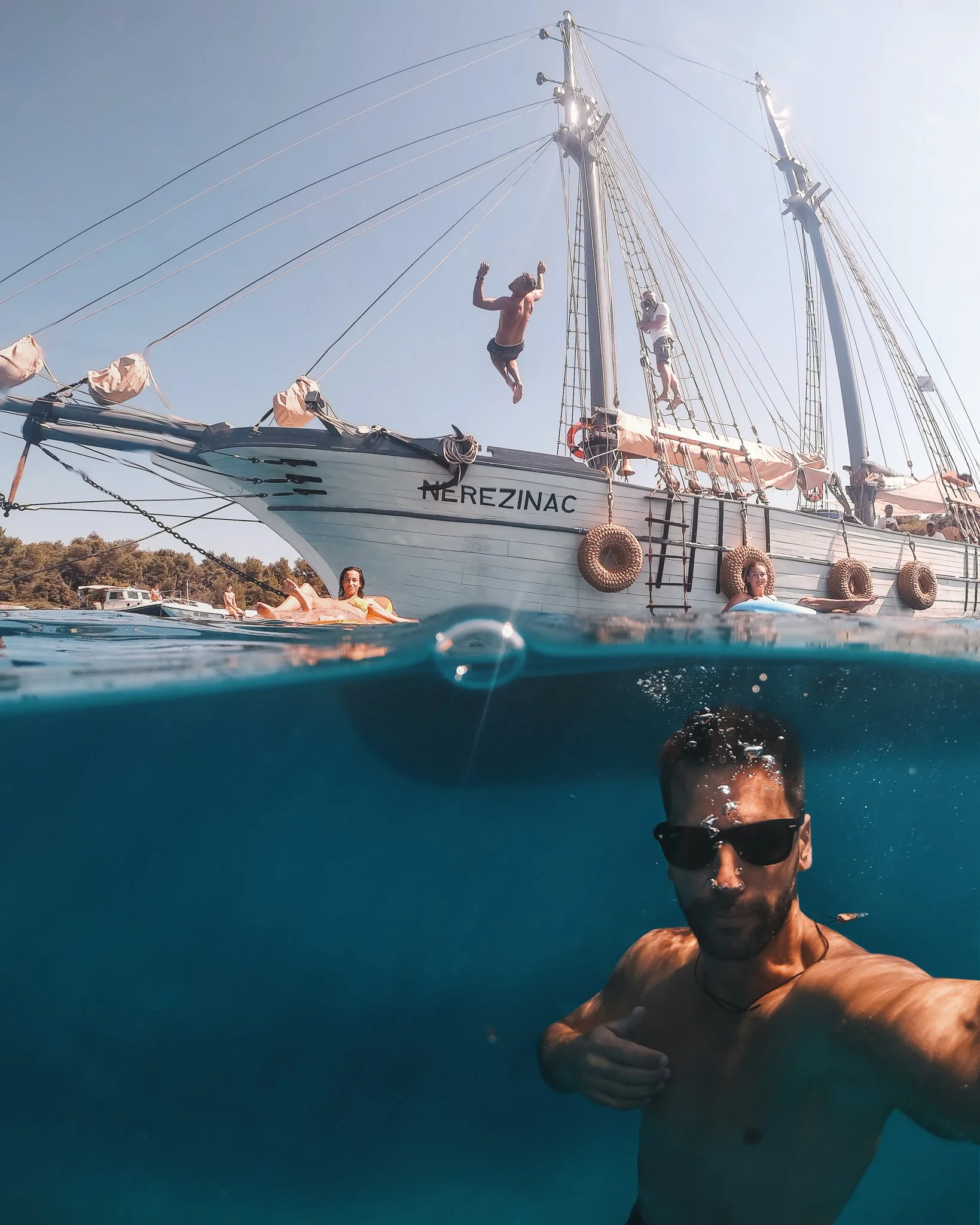 A man is taking a selfie underwater near a boat, with two women floating nearby. The boat has the name 'NEREZINAC' on its hull and is anchored at a dock with other boats in the background. The man is shirtless, wearing sunglasses, and has short dark 