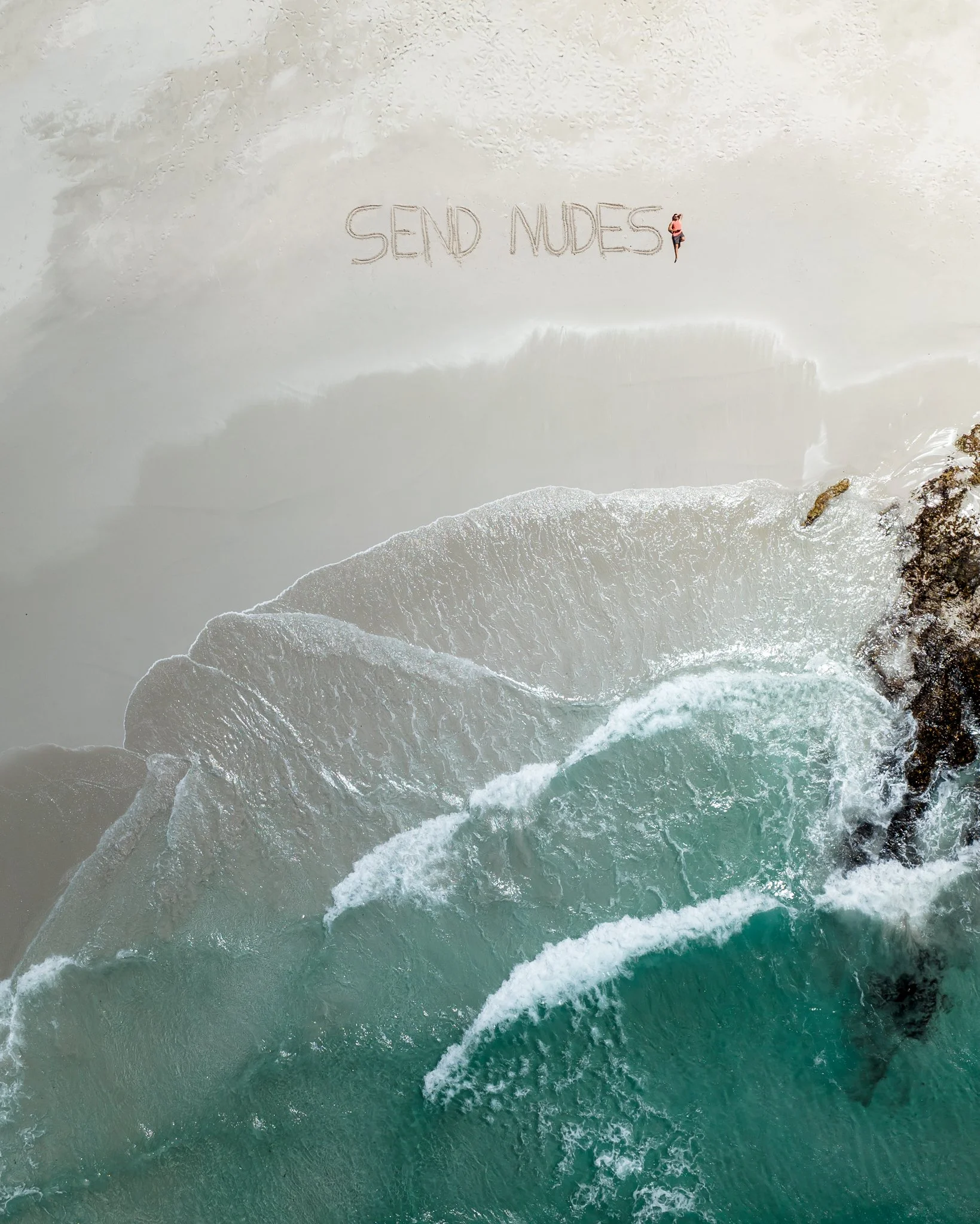 A person sitting on a sandy beach near the ocean, with the words 'SEND NUDES' written in the sand nearby.