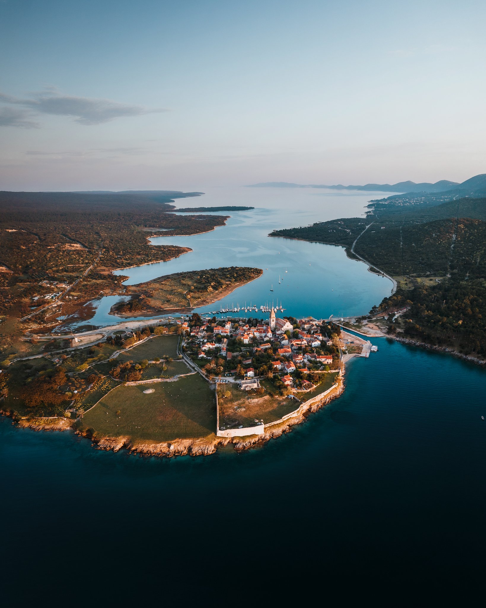Aerial view of a small town on a peninsula surrounded by water, with boats docked at a marina and mountains in the background.