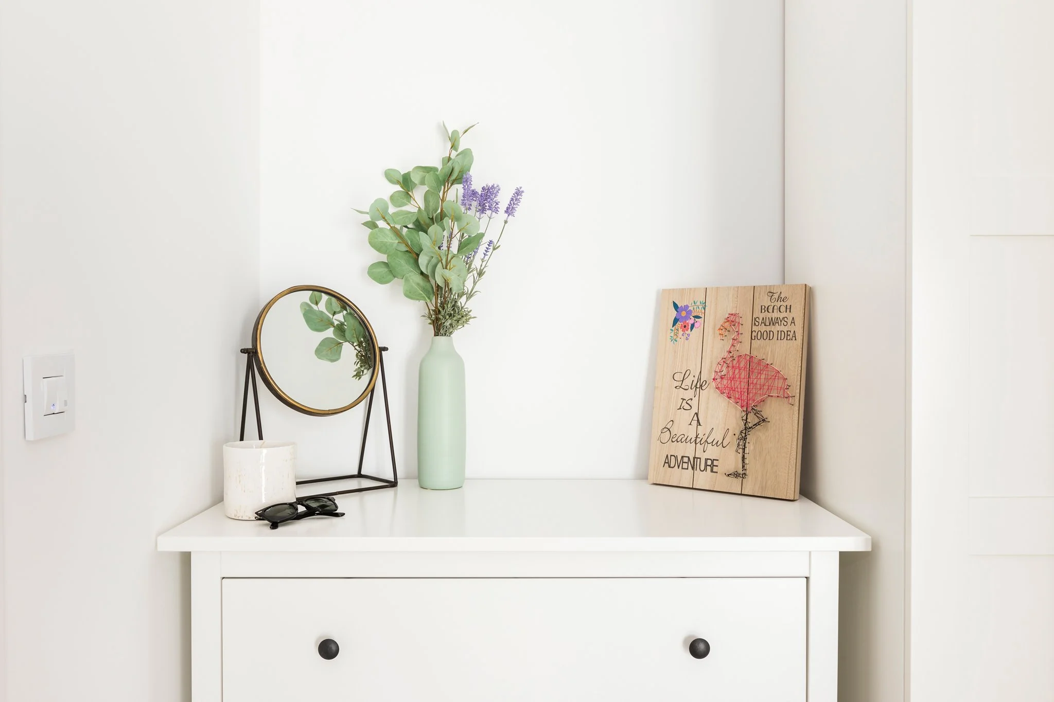 Decorative vanity table with a tall green vase containing eucalyptus and lavender, a small mirror on a black stand, a white textured cup, sunglasses, and a wooden sign leaning against the wall with art and quote about the beach and adventure, set aga