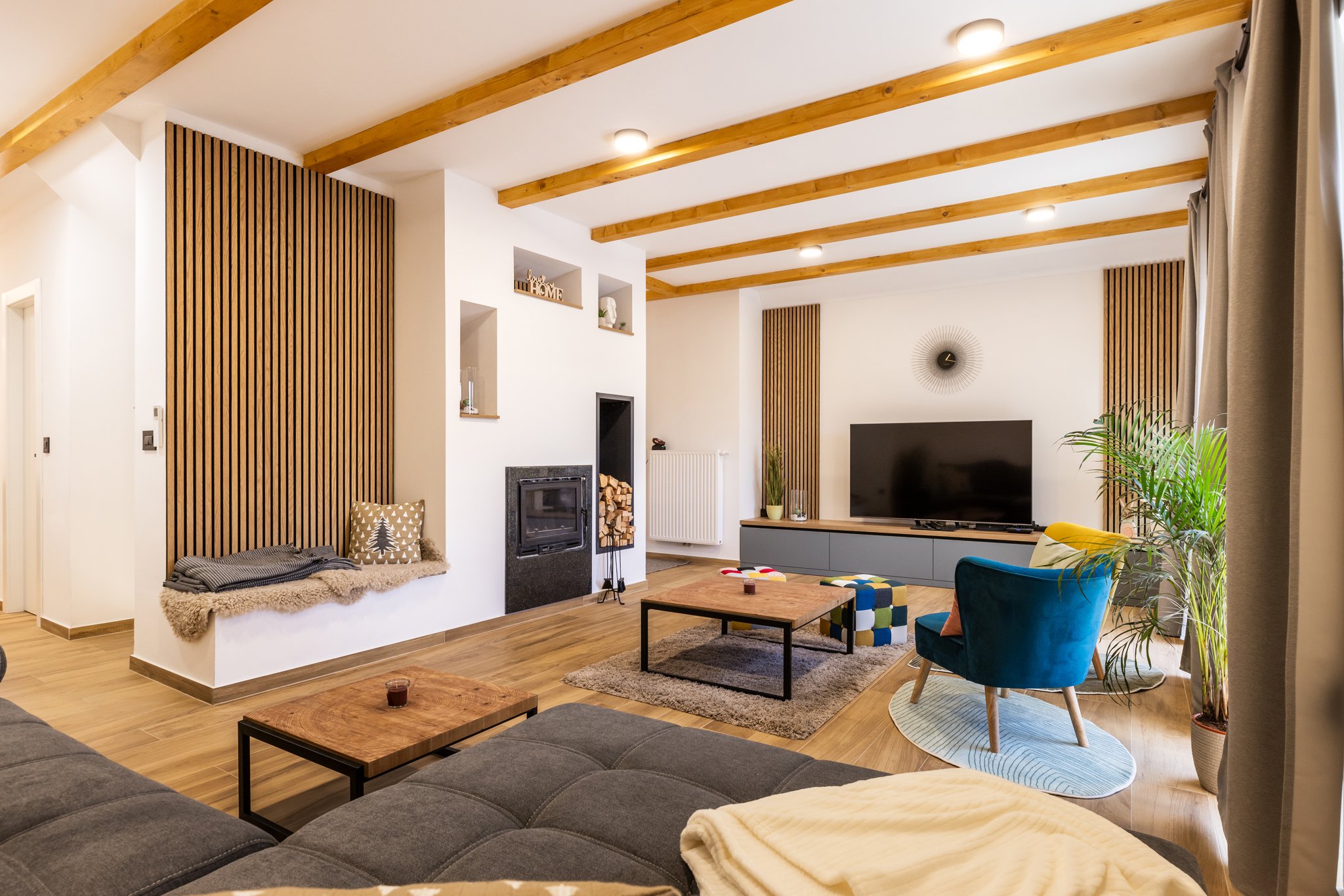 Living room with white walls and wooden ceiling beams, a black fireplace, and a large flat-screen TV on a gray cabinet. Decor includes potted plants, a wall clock, and wall art. Furniture features a gray sectional sofa with throw blankets, a blue arm