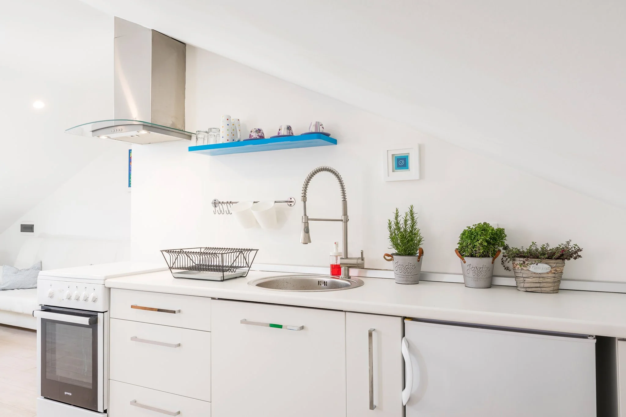A modern white kitchen with a countertop, plants, and a small digital screen on the wall.