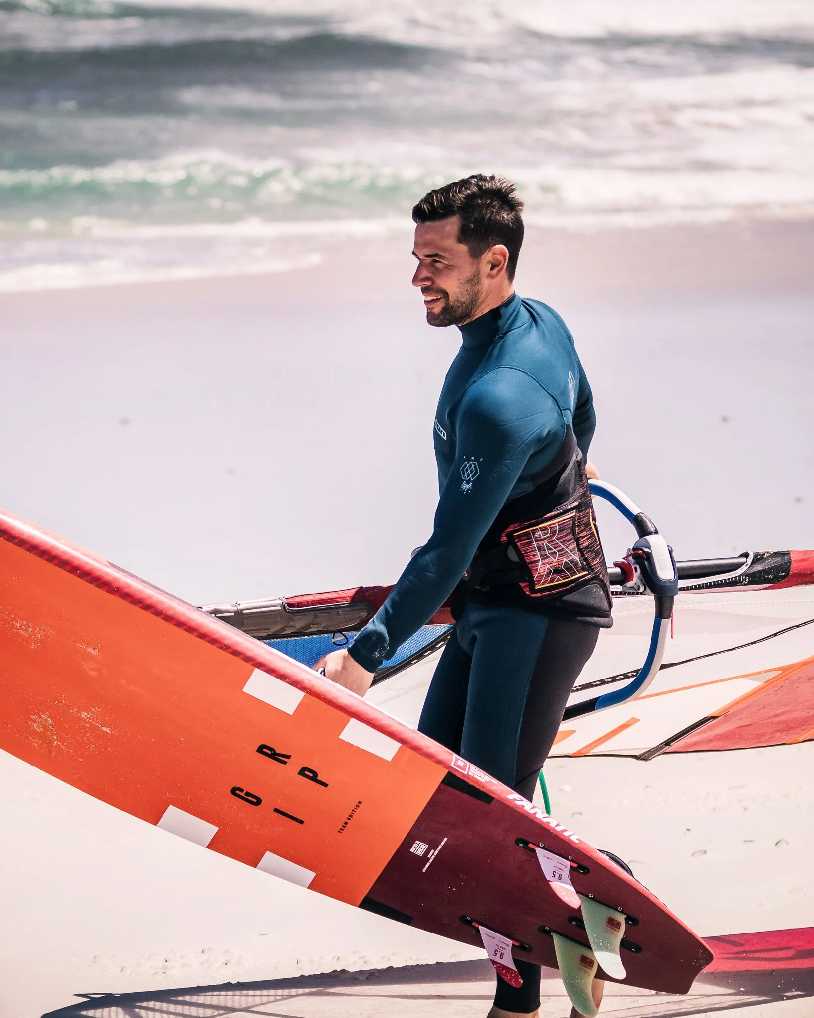 A man dressed in a wettie standing on the beach with multiple surfboards. Abstract ocean water can be seen in the background.