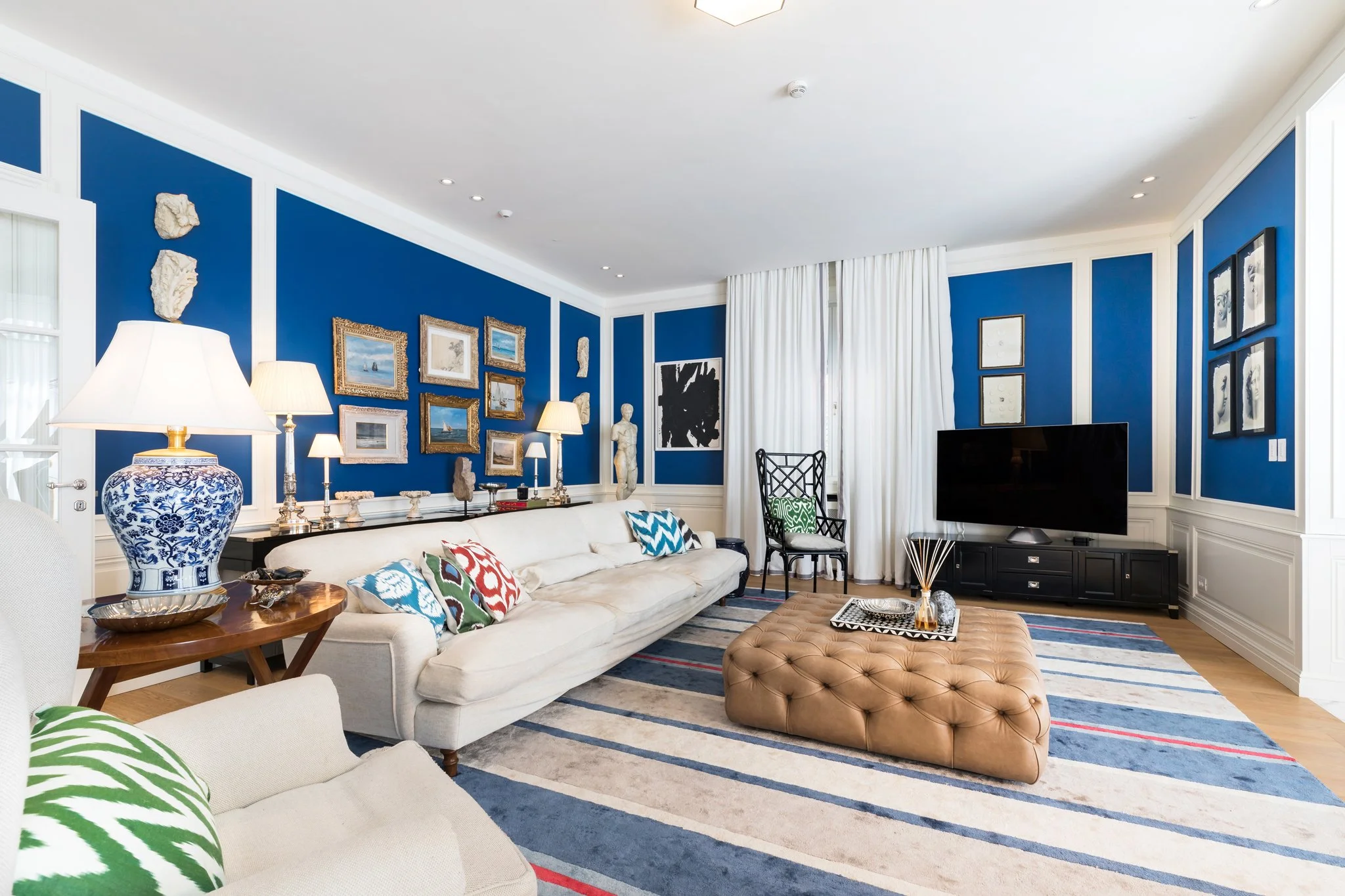 Living room with blue walls decorated with framed artworks and sculptures, white curtains, a white sofa with colorful pillows, a patterned rug, a large blue and white vase on a wooden table, multiple lamps, a flat-screen TV, and an orange tufted otto