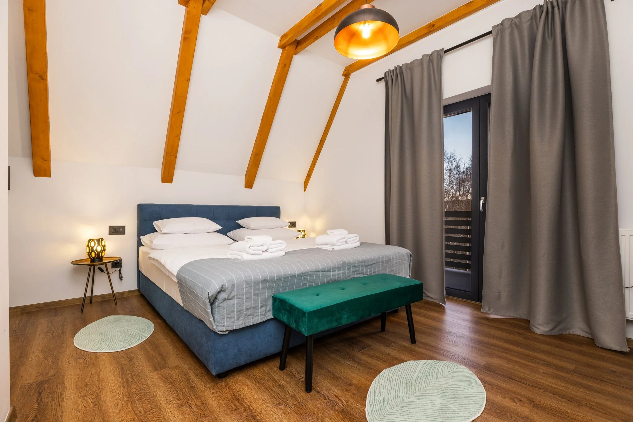 A bedroom with a sloped ceiling featuring exposed wooden beams, a blue upholstered bed with white pillows, and neatly folded towels on top. A small round side table with a decorative lamp is beside the bed. The window is partially covered by grey cur