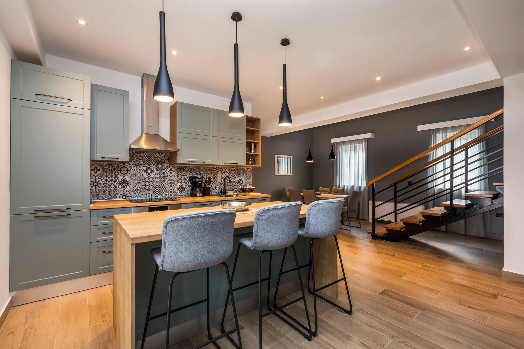 Modern kitchen and dining area with gray cabinets, patterned backsplash, wooden countertop, gray chairs around a wooden dining table, black pendant lights, and a staircase with wooden steps and black railing.