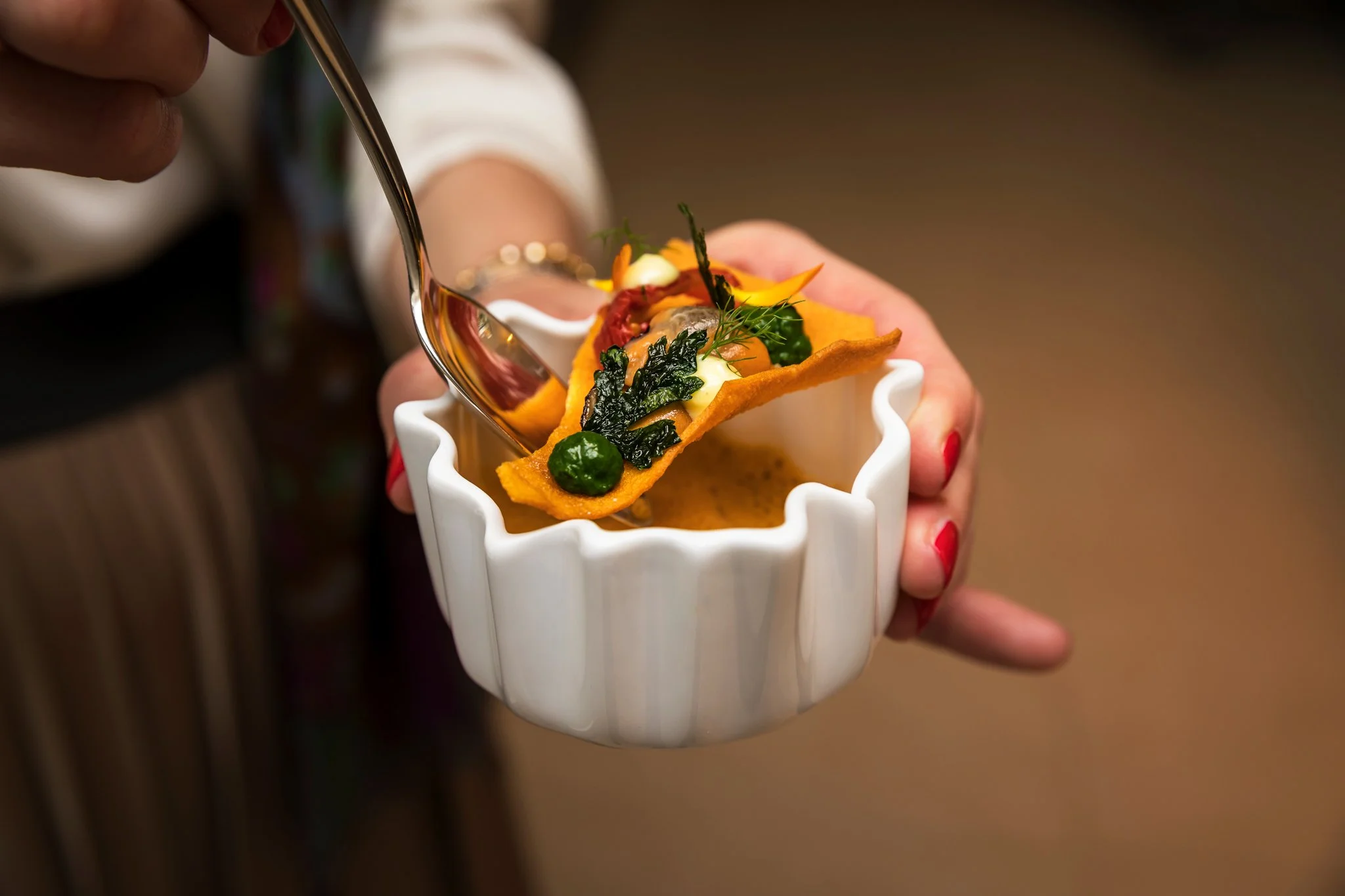 Close-up of a hand holding a white ceramic cup with a savory dish, including colorful vegetables and herbs, being poured with sauce from a spoon.