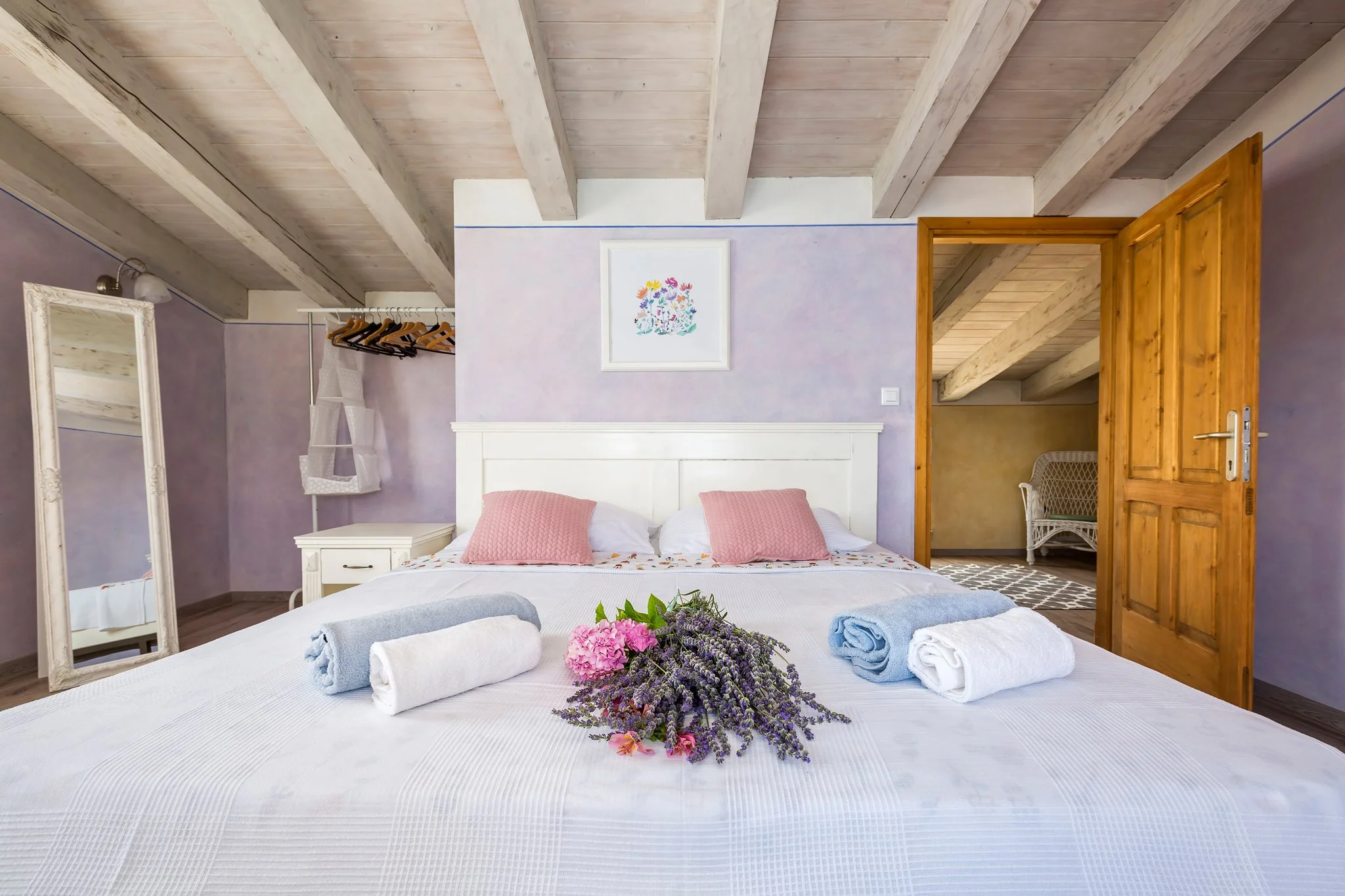 A neatly made bed with white linens, pink pillows, and towels with lavender and pink flowers on top in a room with purple walls, wooden beams on the ceiling, a mirror on the left, and an open wooden door leading to another room.