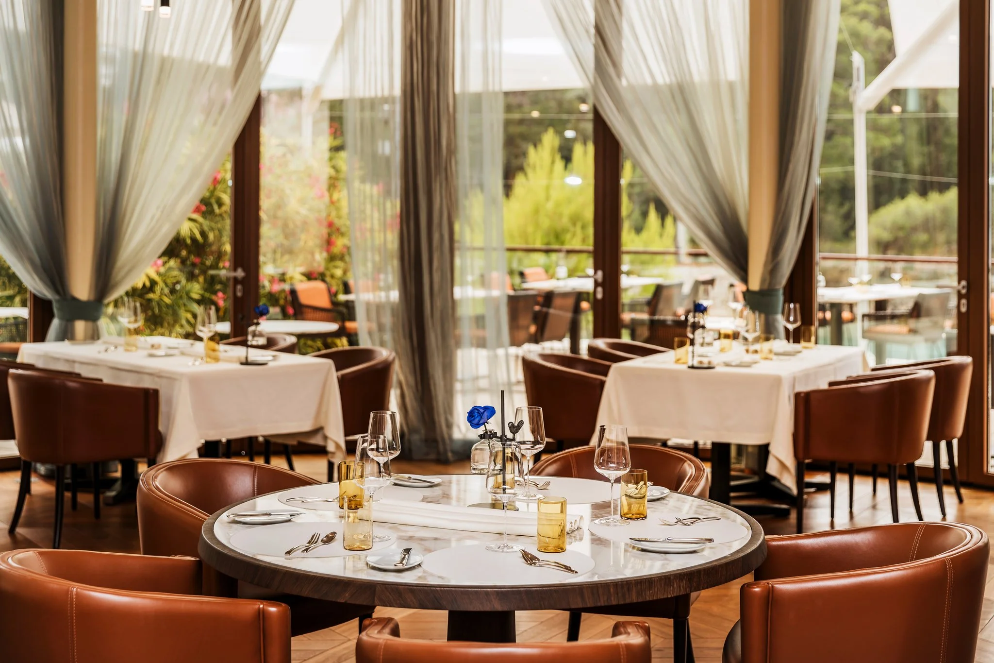Dining room with round tables covered with white tablecloths, set with wine glasses, cutlery, and folded napkins. The room has large windows with sheer curtains, overlooking an outdoor patio with greenery and trees.Alfred Keller in Mali Lošinj, Bouti