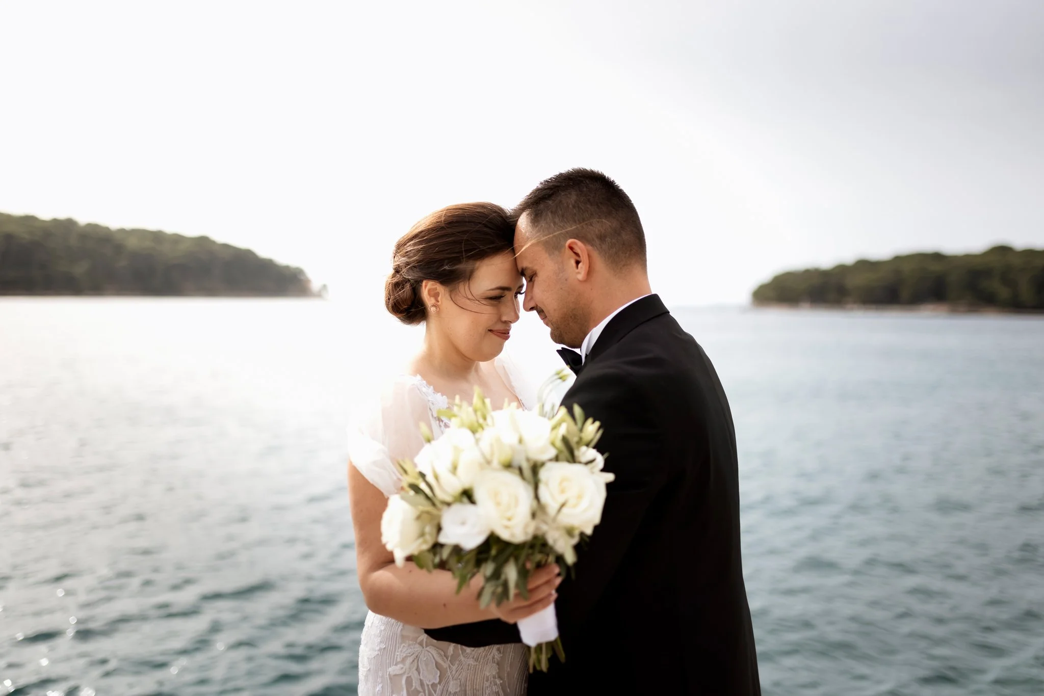 A bride and groom are standing close together, with their foreheads touching, on a body of water with small islands or landmasses and a cloudy sky in the background. The bride is wearing a white dress and holding a bouquet of white flowers, while the