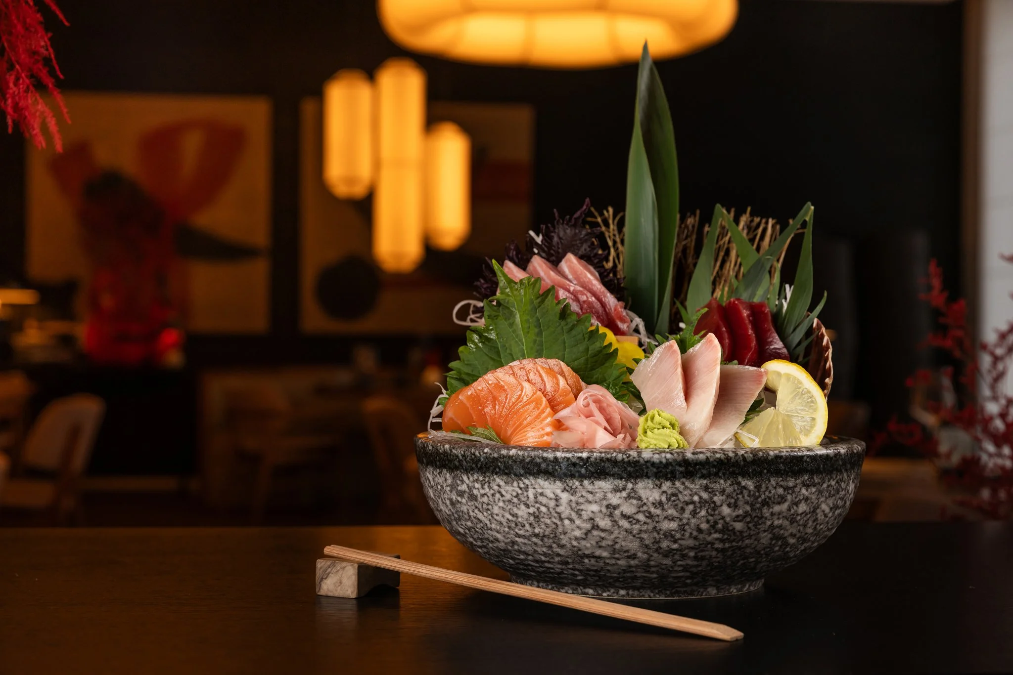 A bowl of assorted sashimi, including salmon, tuna, and other fish, garnished with lemon slices, green leaves, and wasabi, placed on a wooden table with chopsticks beside it. The background features warm lighting and traditional Japanese decor.