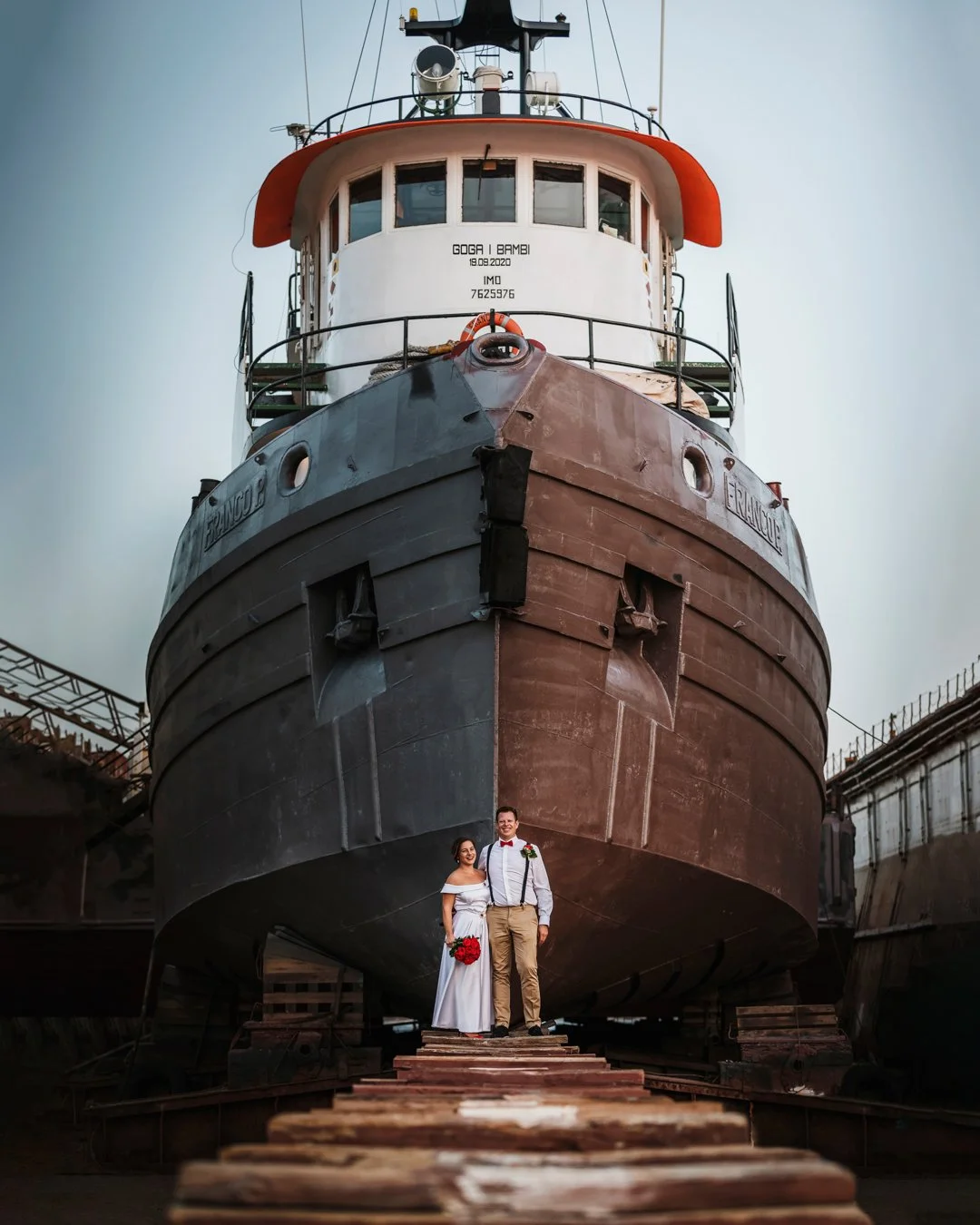 A couple dressed in wedding attire standing under a large ship in a shipyard, with the ship's hull towering above them.