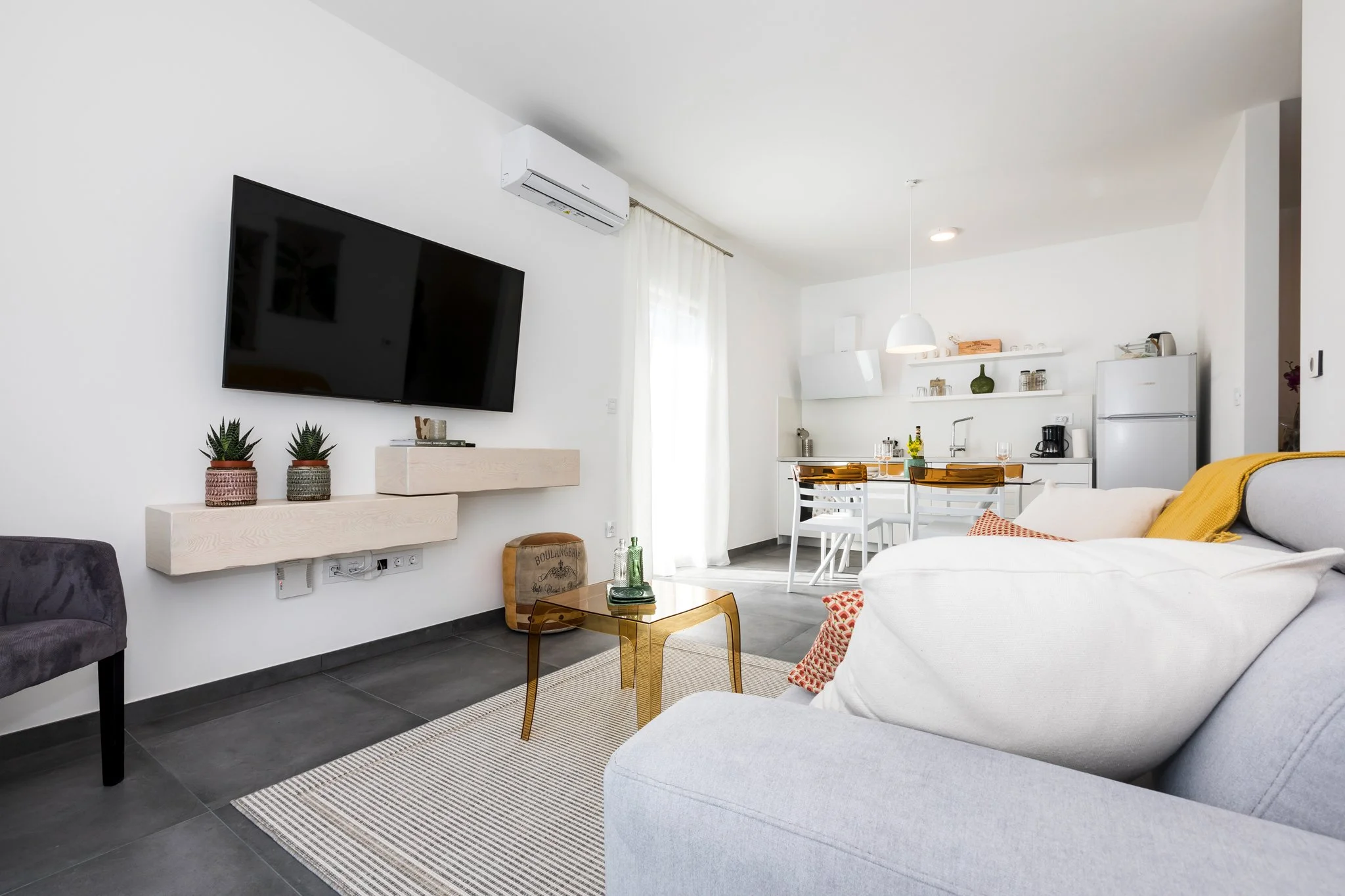 A modern, minimalistic living room with a white couch, gold coffee table, gray armchair, flat-screen TV on a white wall, and a visible kitchen area with white cabinets, a refrigerator, and a dining table with yellow and white chairs.