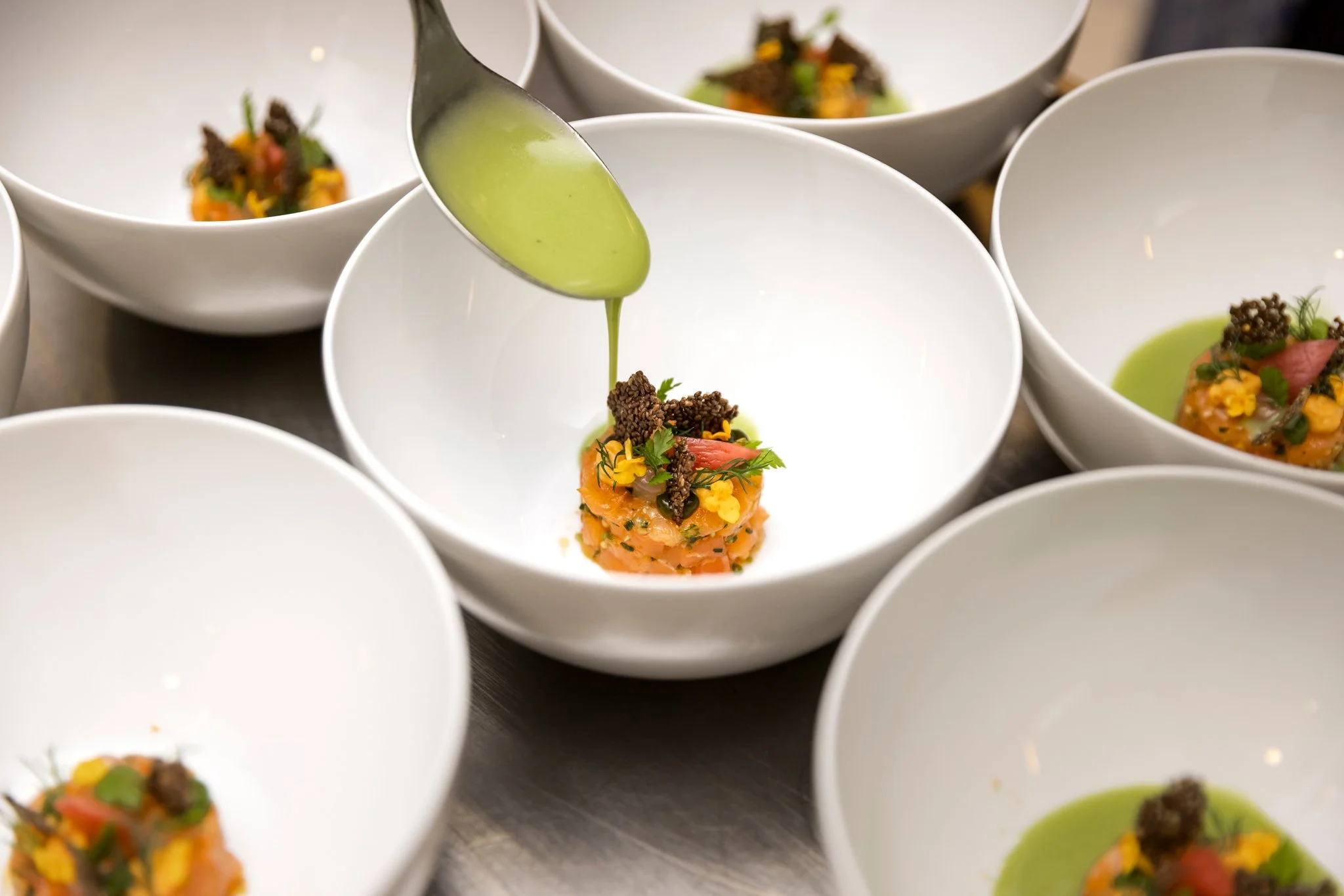 Close-up of several white bowls filled with culinary dishes, one being spooned with a green sauce, and some containing a colorful vegetable medley with herbs and garnishes.
