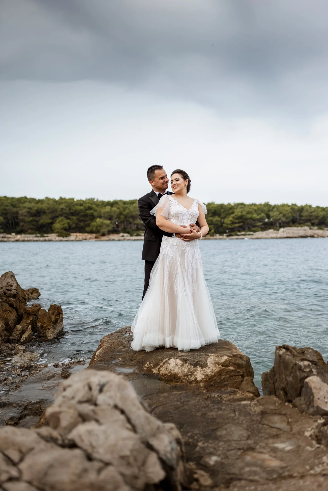 A couple in wedding attire standing on large rocks by the water, with a tree-lined shoreline and cloudy sky in the background.