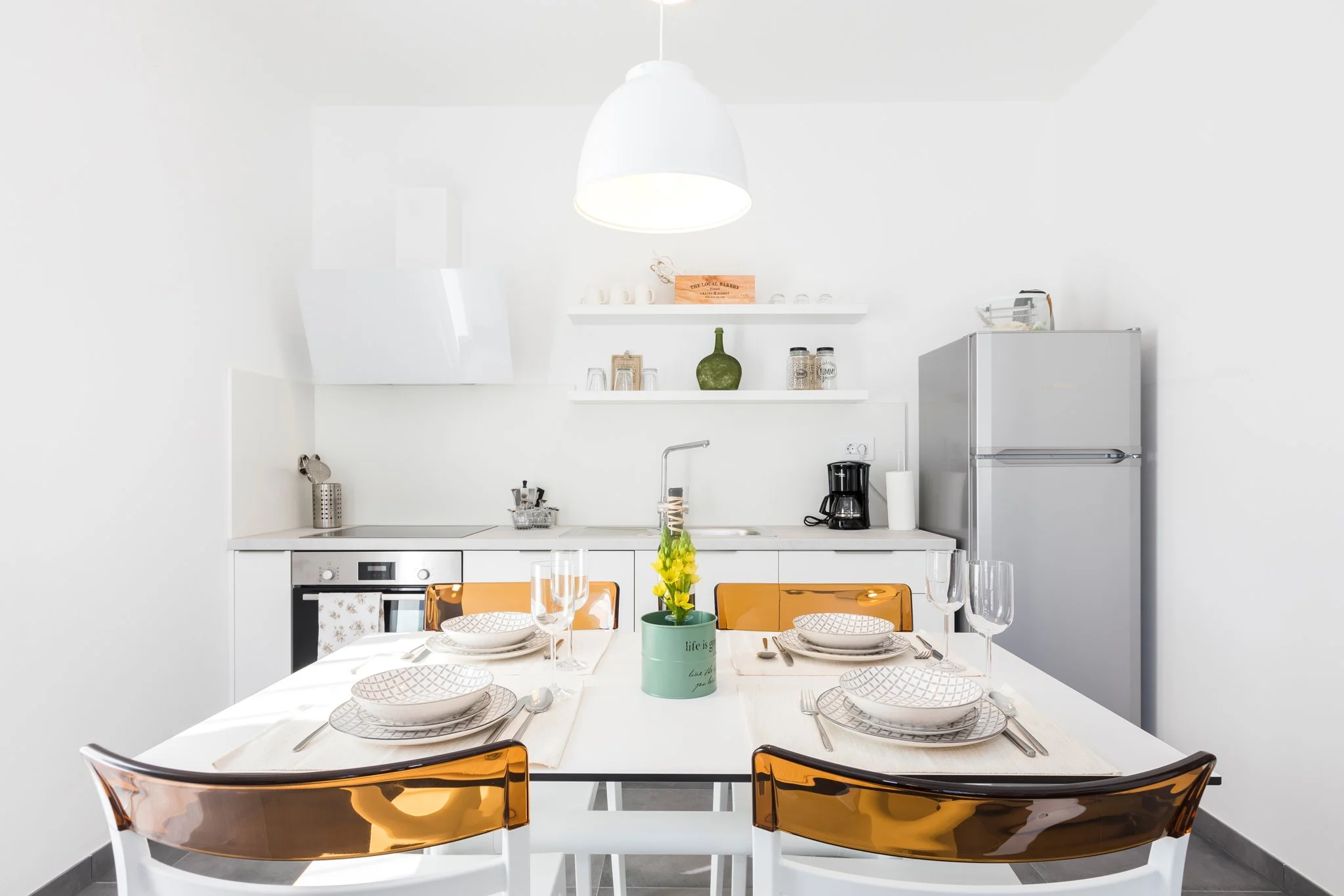 Modern, white kitchen with a dining table set for four people, including white plates, glasses, and silverware, with a plant in a green pot in the center.