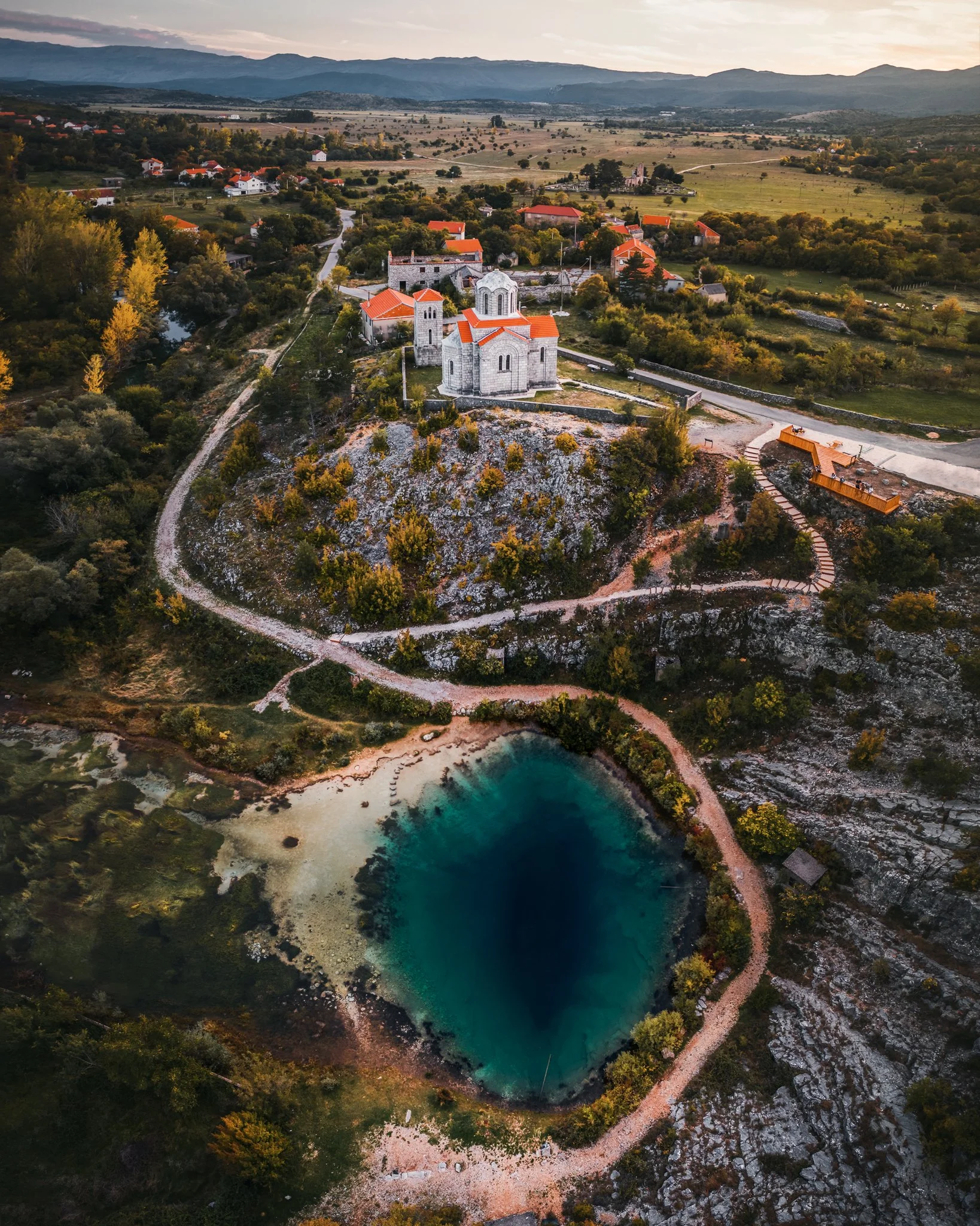 Aerial view of a small village with a historic stone church on a hilltop, surrounded by trees and hiking paths, with a lake in the foreground and mountains in the distance during sunset.