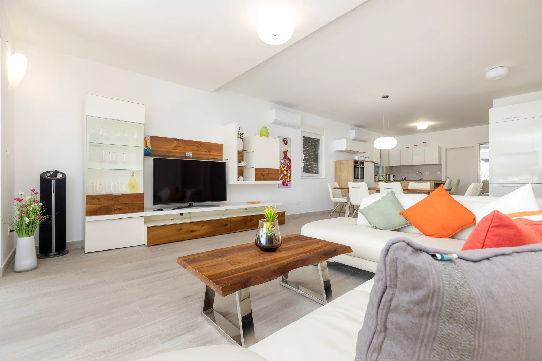 Modern open-concept living room and kitchen with white walls, wooden accents, colorful cushions, a flat-screen TV, and minimal decor.