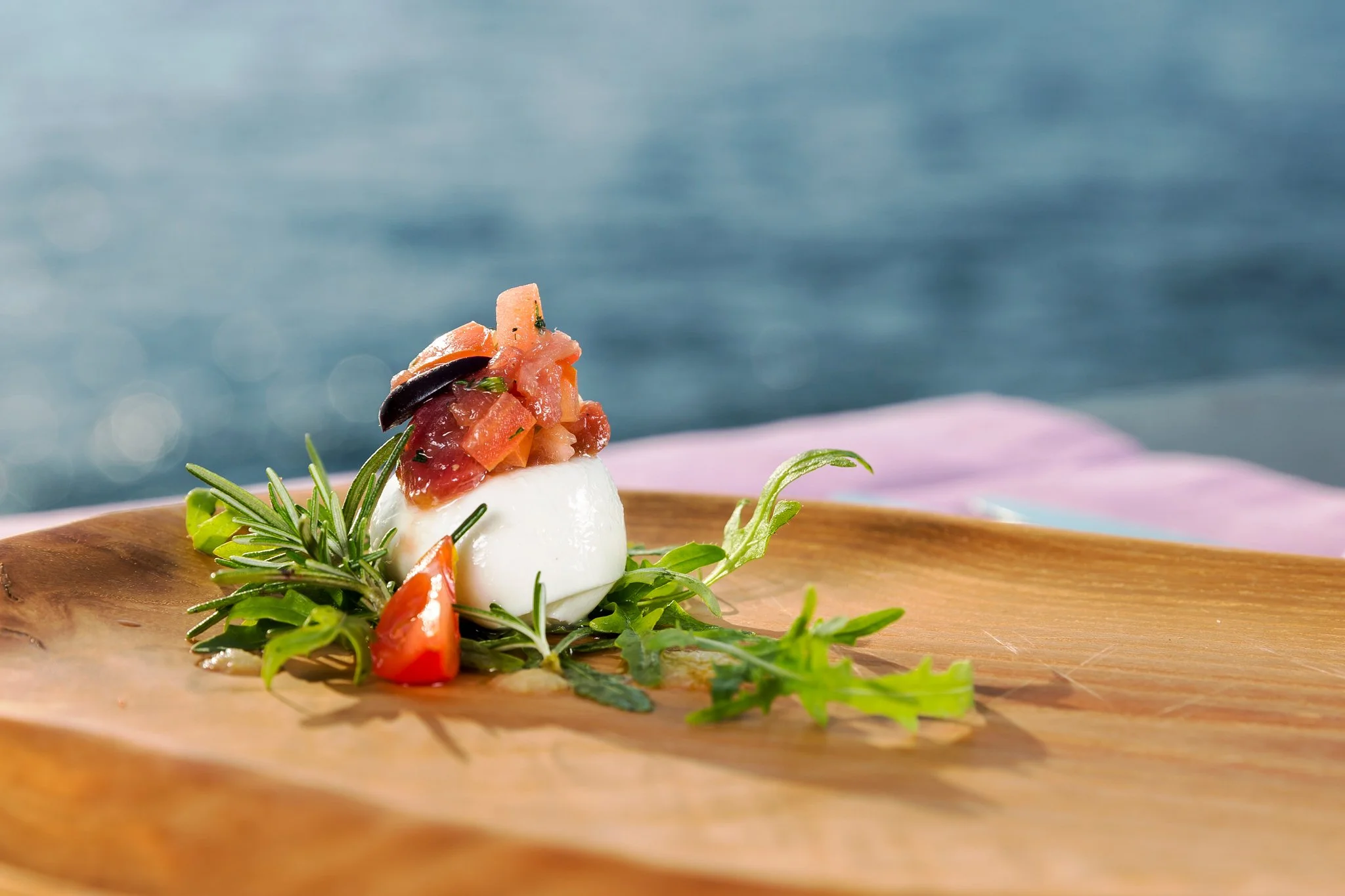 Poached egg topped with diced tomatoes, black olives, and chopped herbs, garnished with fresh greens, on a wooden plate with a blurred water background.