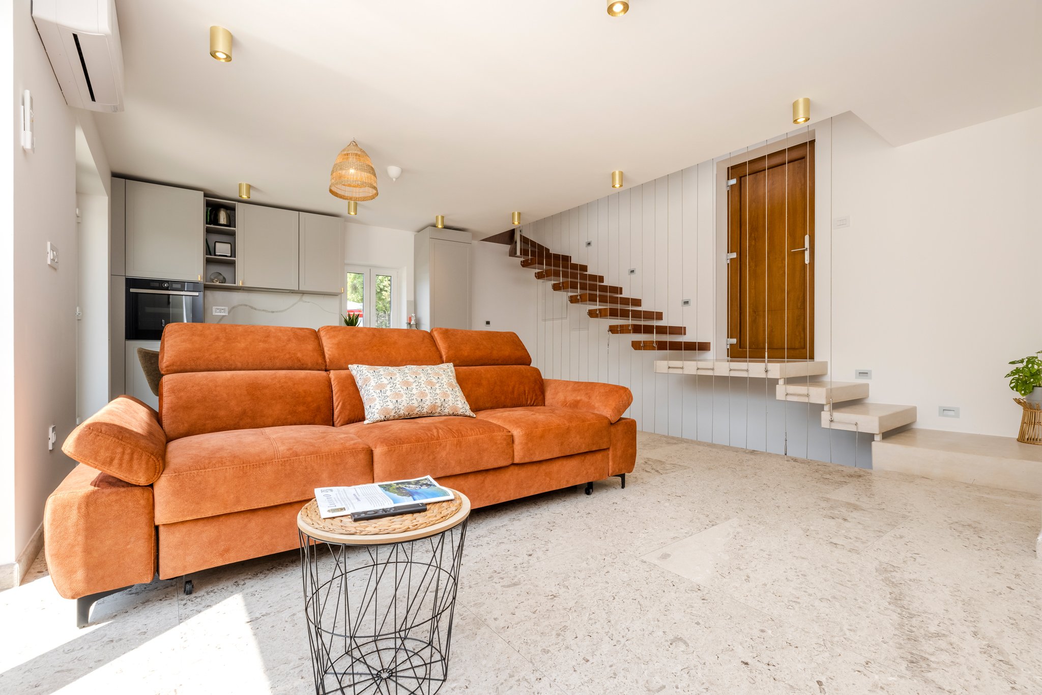 Living room with a rust-colored sofa, a small round side table with a magazine and remote, a staircase with floating wooden steps, and a kitchen area in the background.
