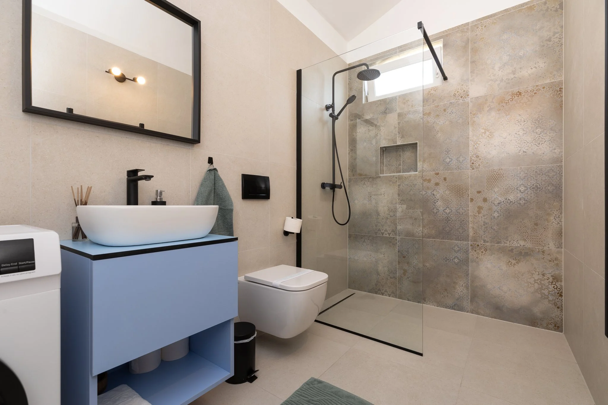 Modern bathroom with a walk-in shower featuring patterned tile walls, a floating toilet, a blue vanity with a white vessel sink, a black faucet, and a large mirror. There is a small window above the shower, a green towel hanging on a hook, and a blac