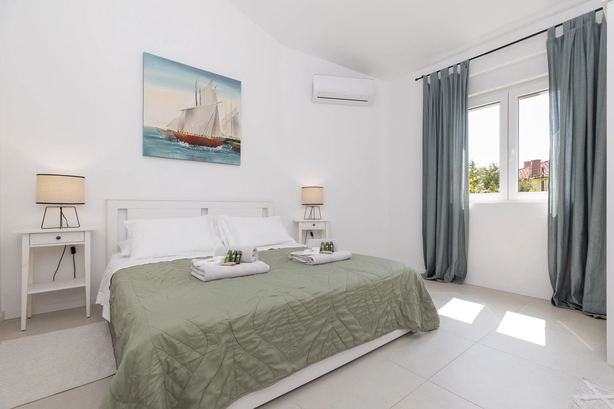 A bright bedroom with white walls, a white bed with green bedding, and two nightstands with beige lamps. A painting of a sailboat hangs above the bed. There are two windows with blue curtains, and an air conditioning unit on the wall.