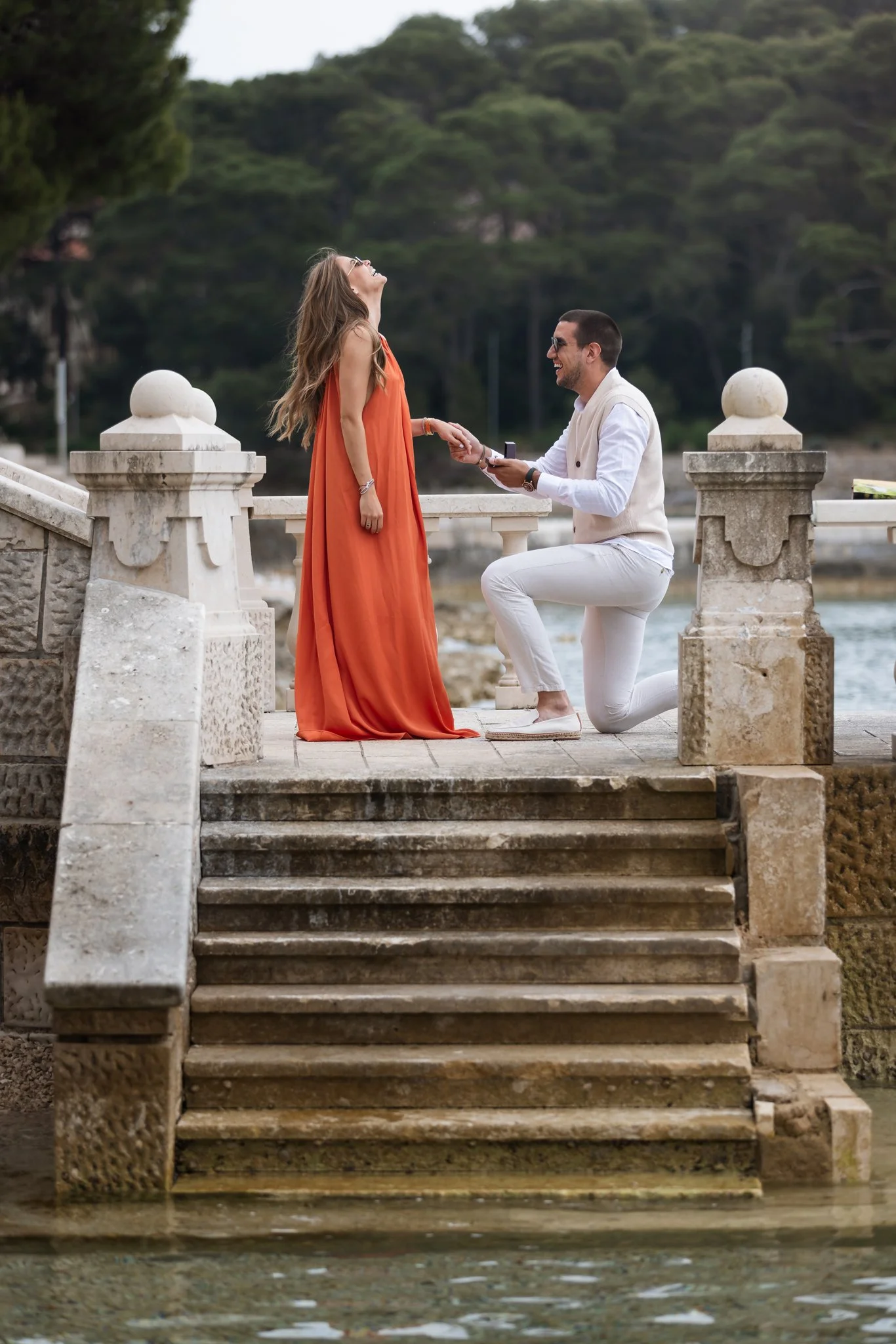 A man kneeling on one knee, proposing marriage to a woman on a stone staircase by the water, with a scenic green forest in the background. The woman is wearing an orange dress and the man is dressed in white.