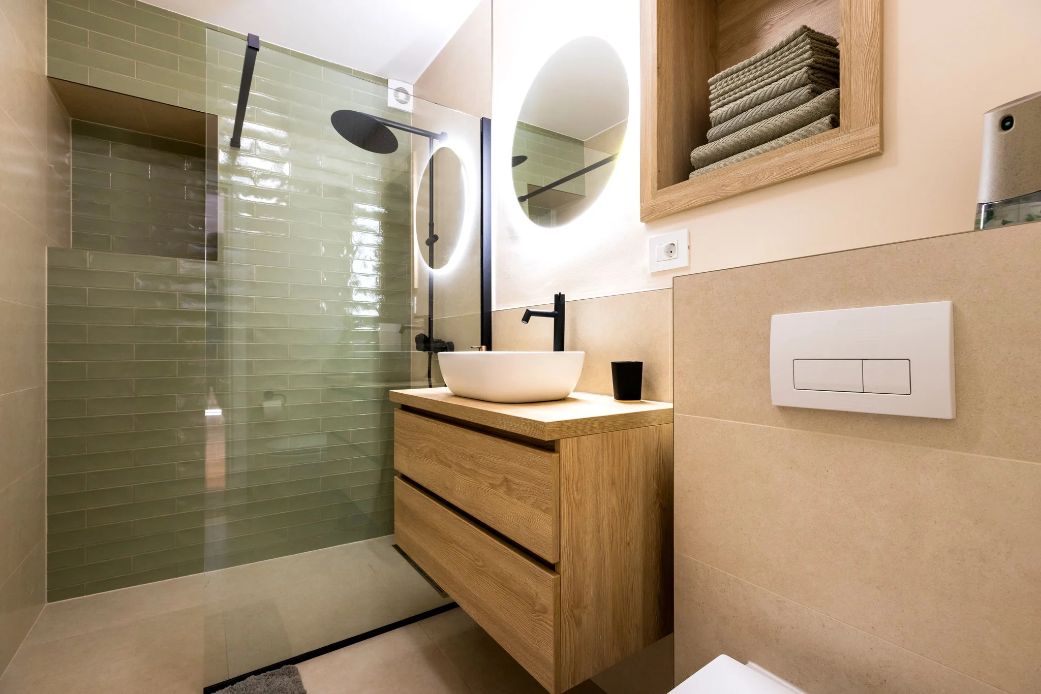 Modern bathroom with a walk-in shower with green subway tiles, a round mirror with back lighting, a black faucet, a wooden vanity with a round vessel sink, a black cup, a wooden shelf with folded towels, and a wall-mounted toilet.
