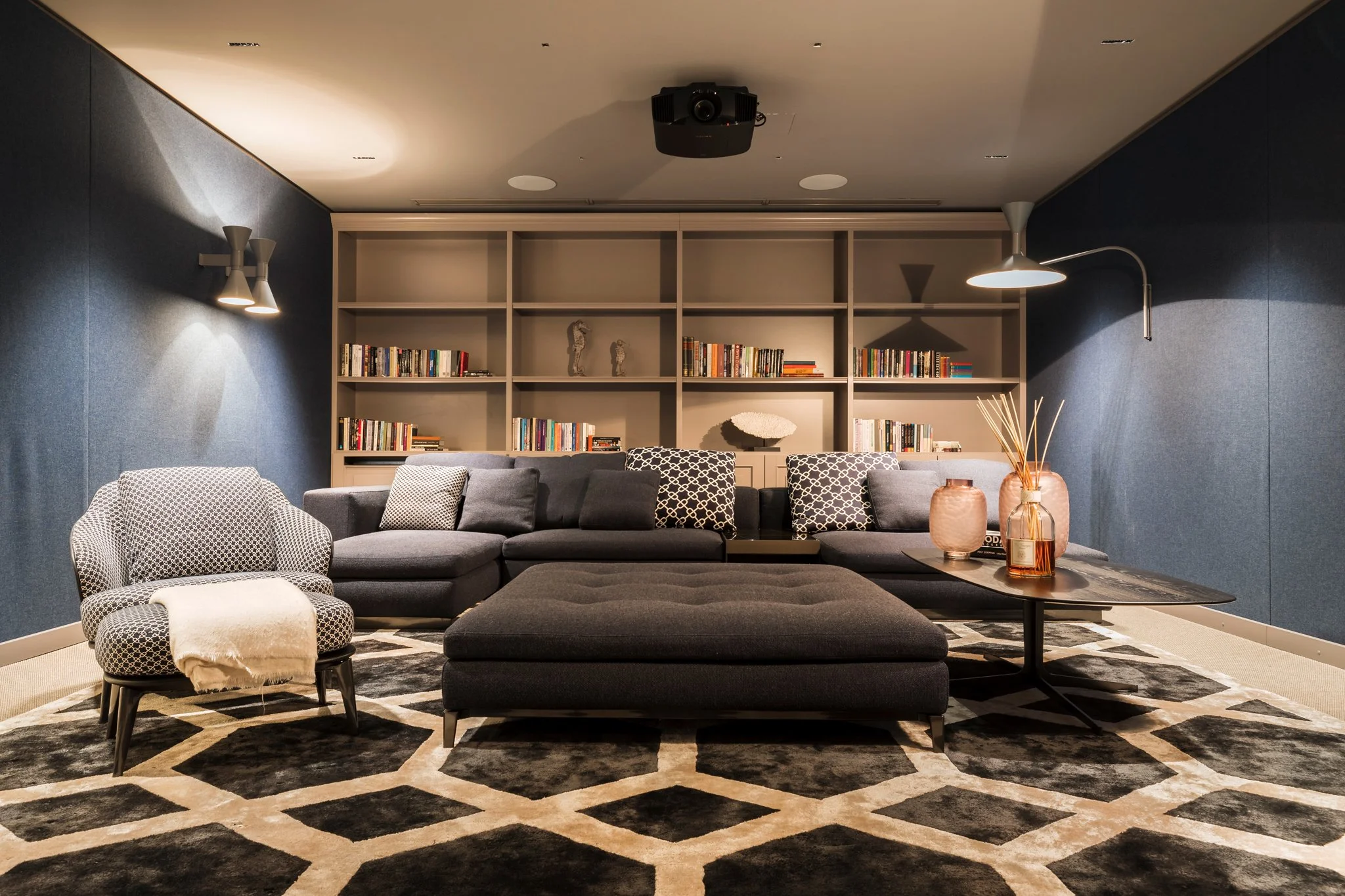 A modern living room with a gray sectional sofa, patterned armchair, decorative vases on a coffee table, bookshelves on the back wall, and wall-mounted lamps, with a patterned area rug and ceiling-mounted projector.
