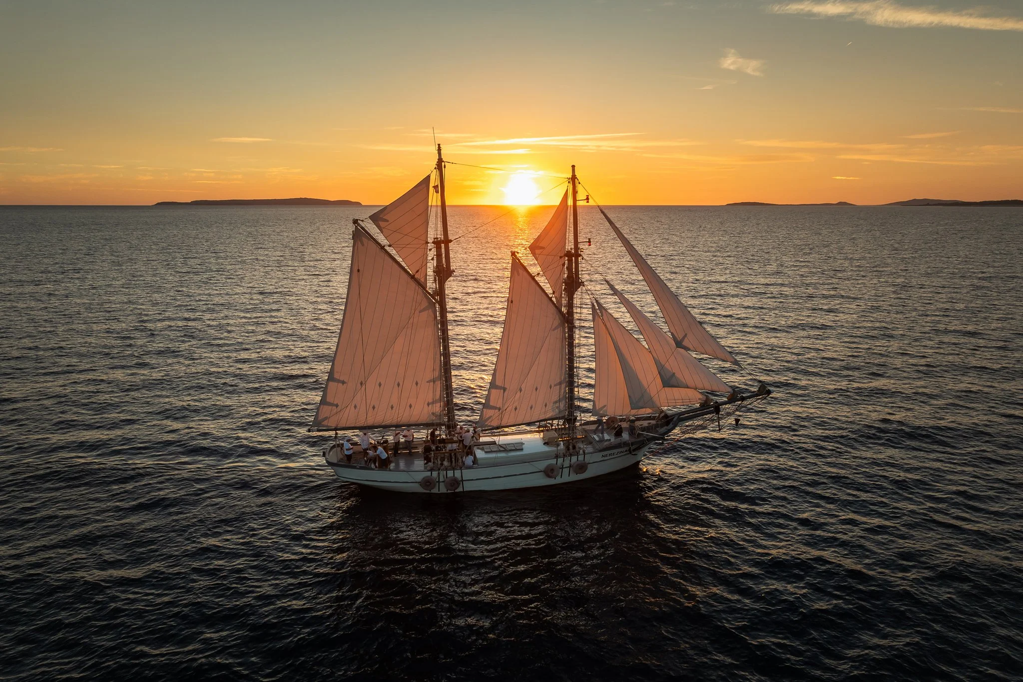 A sailboat with multiple sails sailing on calm water at sunset, with a distant horizon and small islands or landforms.