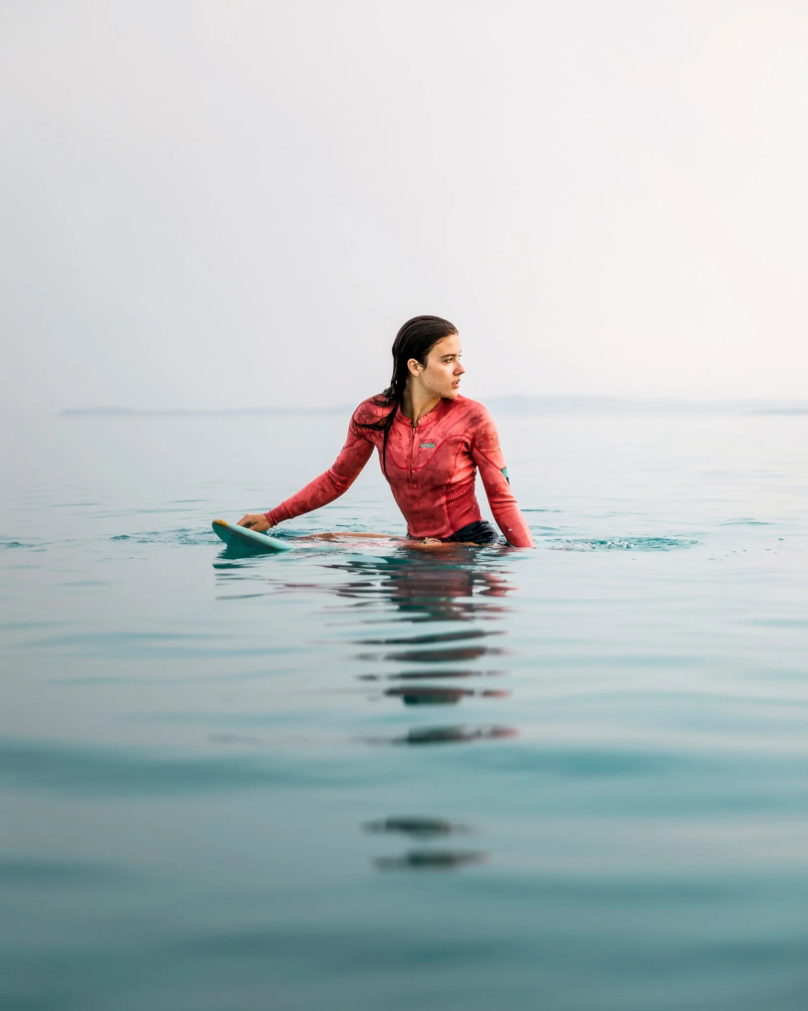 A woman in a red long-sleeved athletic top sits in calm water with a paddle board, looking to her right, with a foggy horizon in the background.