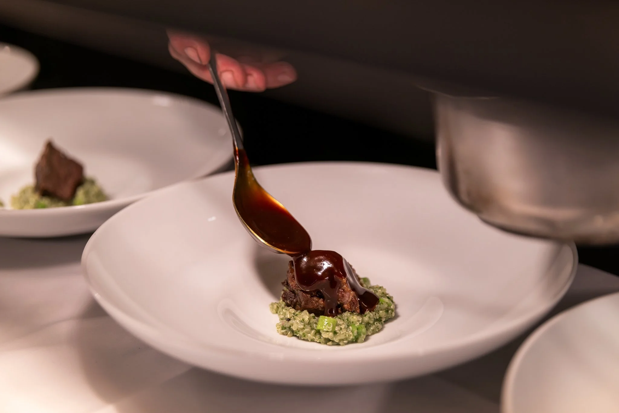 A chef pouring a dark sauce over a plated meat dish on a white plate.