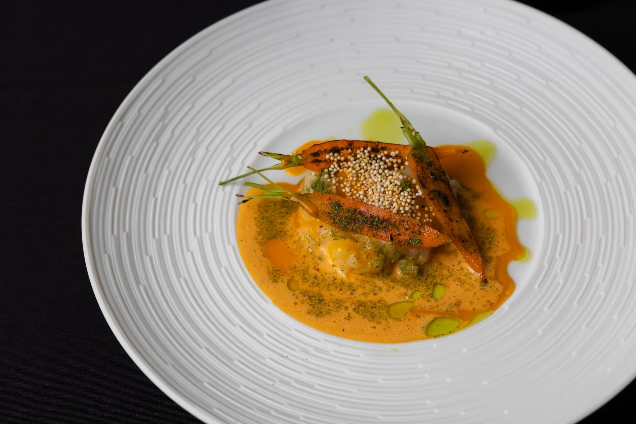 A white bowl with orange sauce, garnished with grilled carrots, microgreens, and white seeds, with a black background.