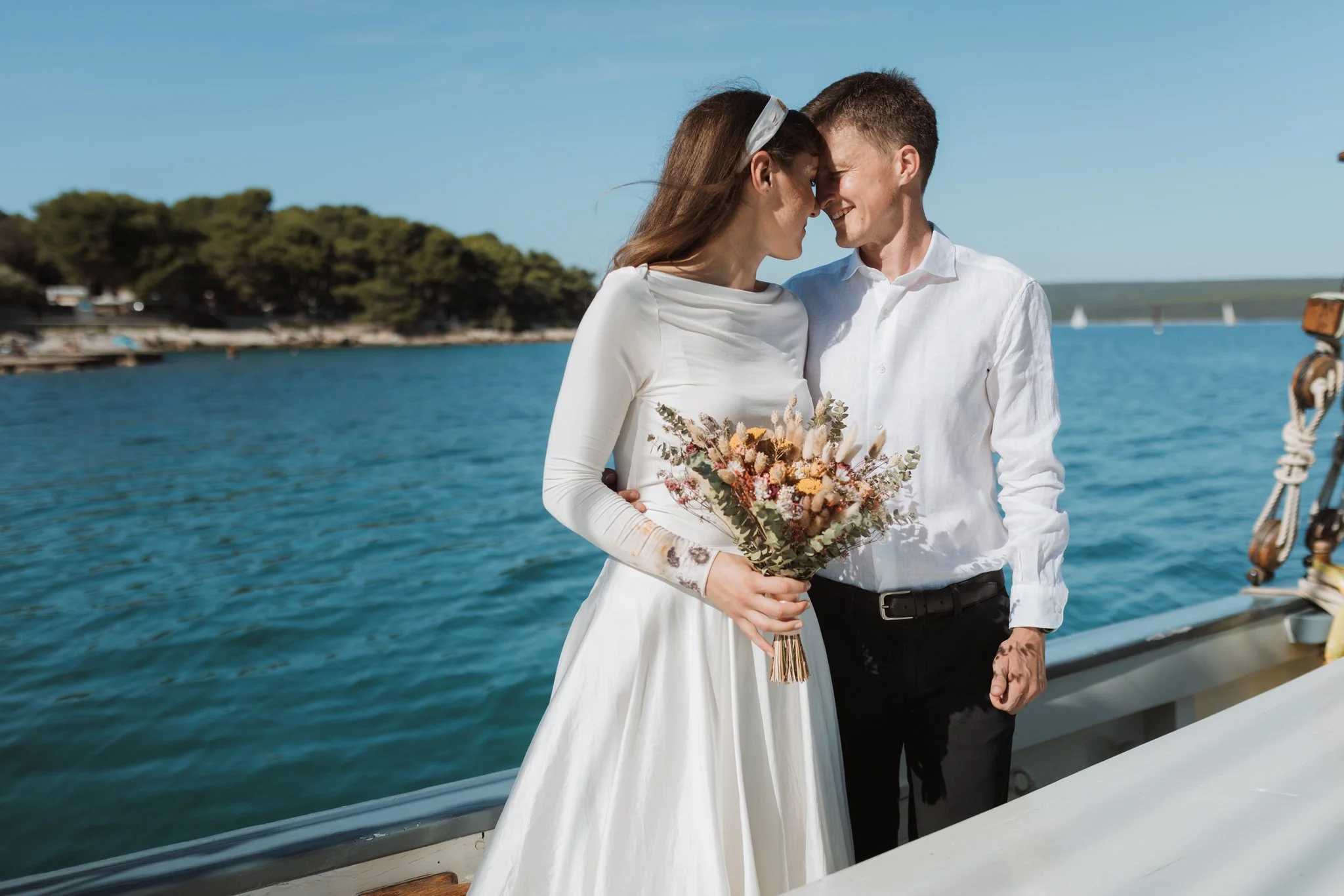 A couple in wedding attire stands closely on a boat dock, touching foreheads, with a woman holding a bouquet of flowers, near a body of water with sailboats in the background, under a blue sky.