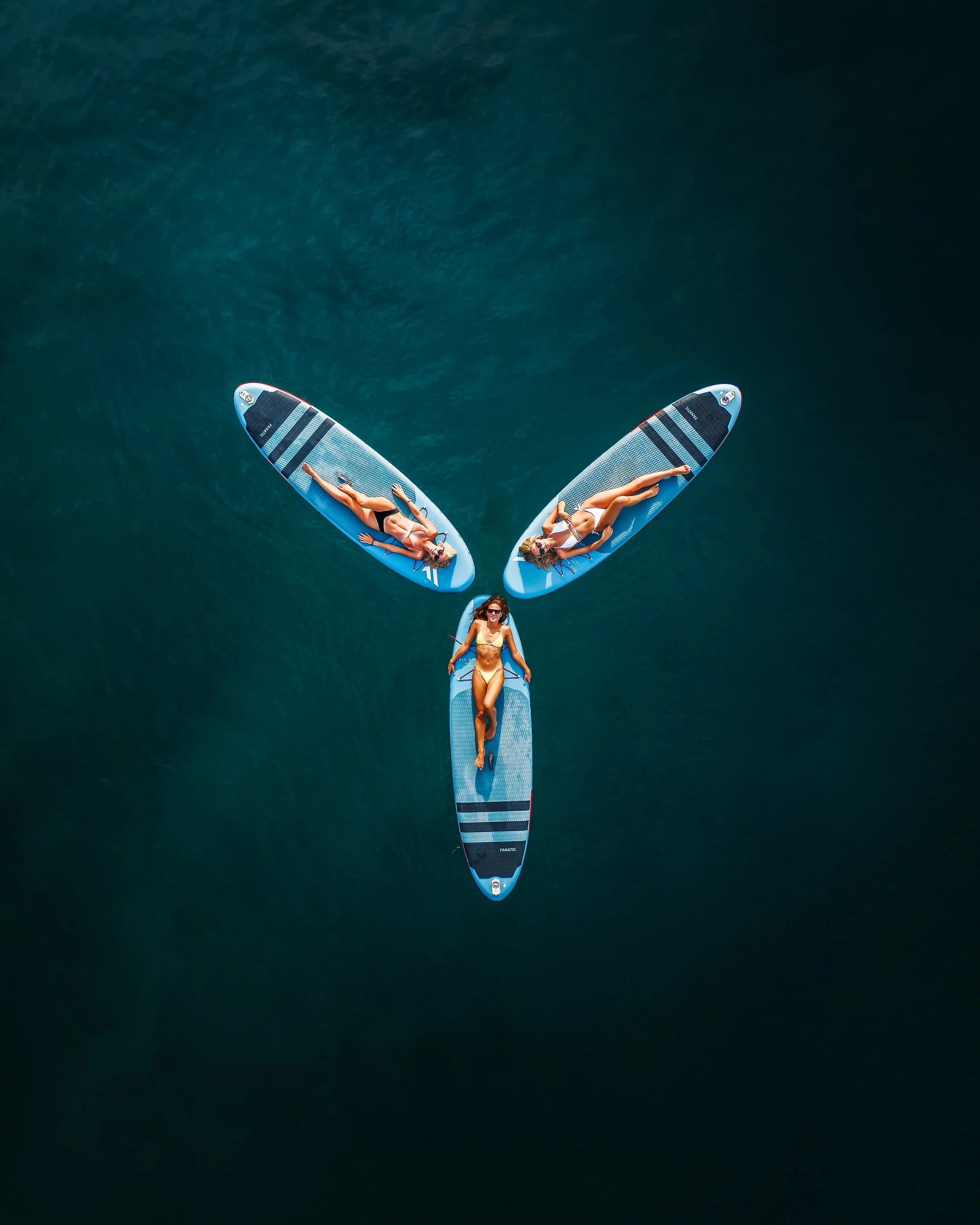 Two women lying on paddleboards in the water, viewed from above, with a third woman standing on the paddleboard at the bottom of the image, all in swimsuits.