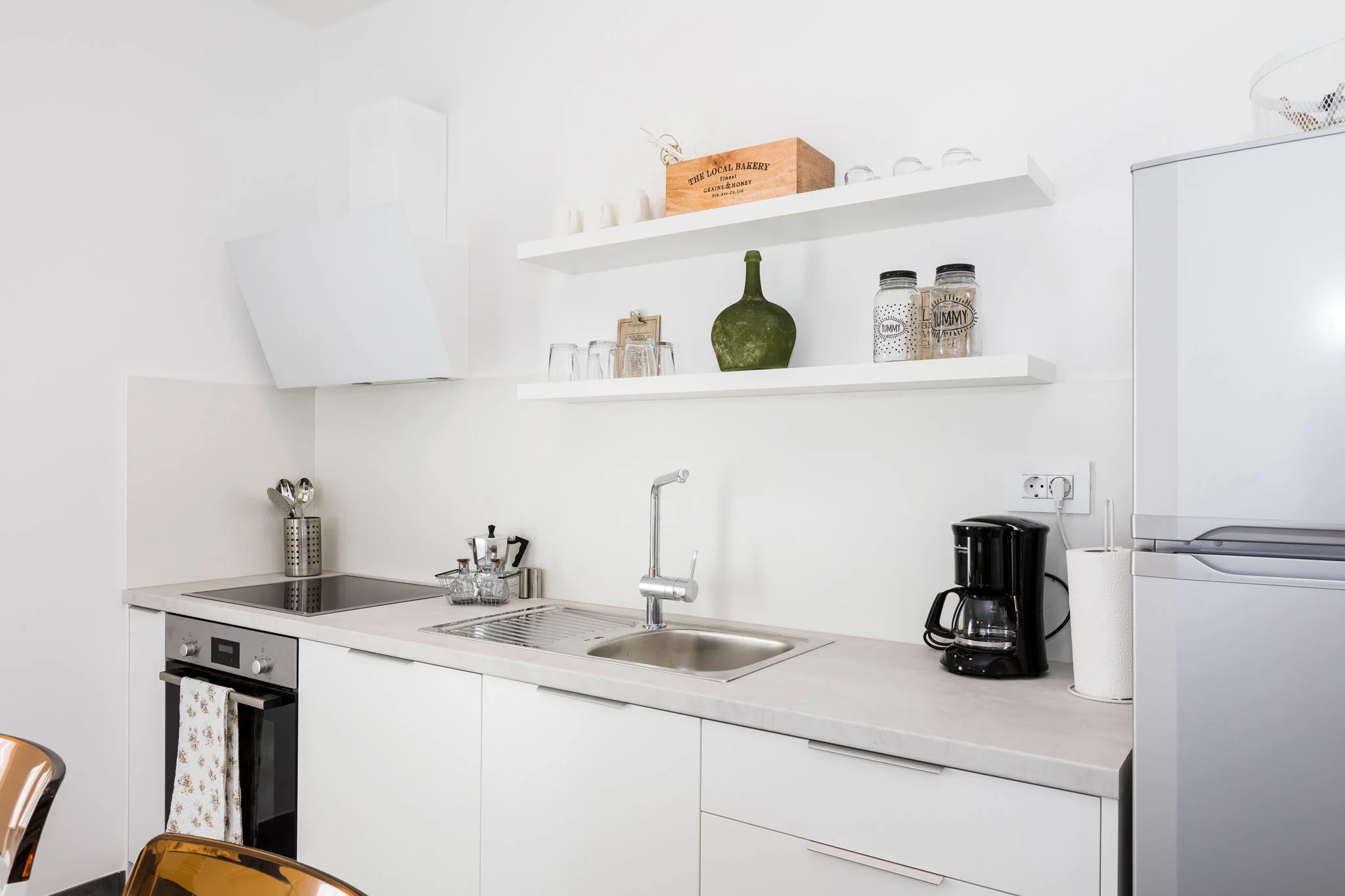 A modern white kitchen with open shelving, a sink, a stovetop, a coffee maker, paper towel roll, and various jars and decorative items on the shelves.