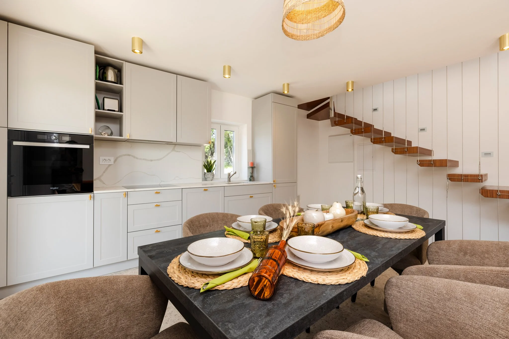 Modern kitchen and dining area with white cabinets, wooden open shelving, a black dining table set with white bowls, green glasses, and a decorated centerpiece. Features a staircase with wooden steps and hanging metal rods, a window with plants, and 